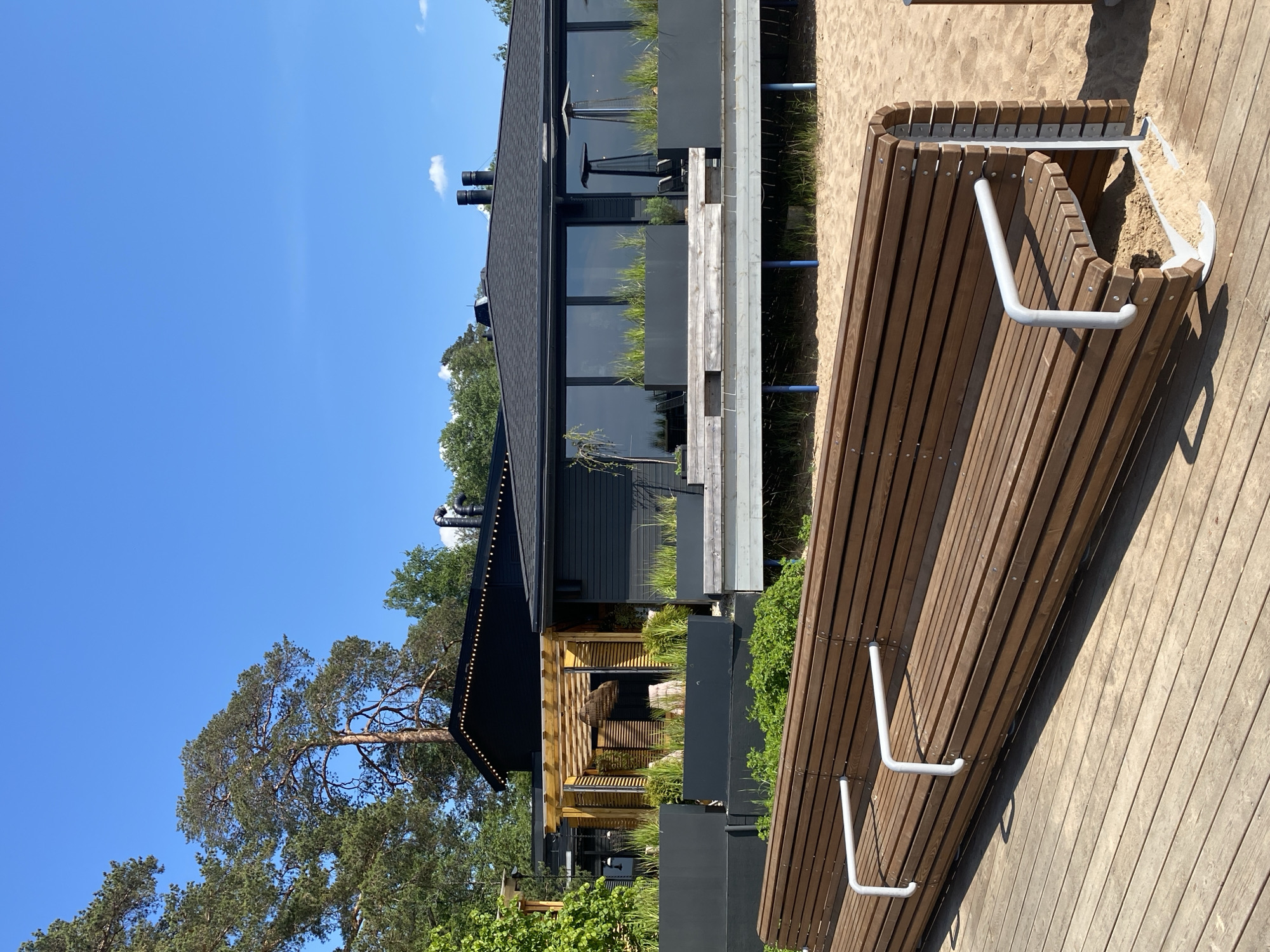 This media file captures a modern outdoor scene in Komarovo, Russia, under a clear, bright blue sky with a few sparse clouds. The image is dominated by contemporary architectural and landscaping elements.
In the foreground and midground, a long, curved wooden bench with multiple slats and white metal handles or armrests is prominent. It is situated on a surface that transitions between a wooden deck or walkway and patches of fine sand.
To the left, a sleek, dark-colored building with a black roof features large, reflective windows that stretch along its facade. Planter boxes filled with green foliage, possibly grasses, are integrated below the windows. Two dark, cylindrical chimney-like structures are visible on the roof. Further back on the left side of the building, a section is covered by a wooden pergola structure, with some visible outdoor furniture underneath.
Green trees, including a large pine tree with distinctive bark, are visible behind and around the building, providing a natural backdrop. The strong, direct sunlight indicates it is daytime, likely late morning or early afternoon, casting distinct shadows across the wooden surfaces and sand. No people are present in the image, contributing to a tranquil and serene atmosphere. There is no visible text.