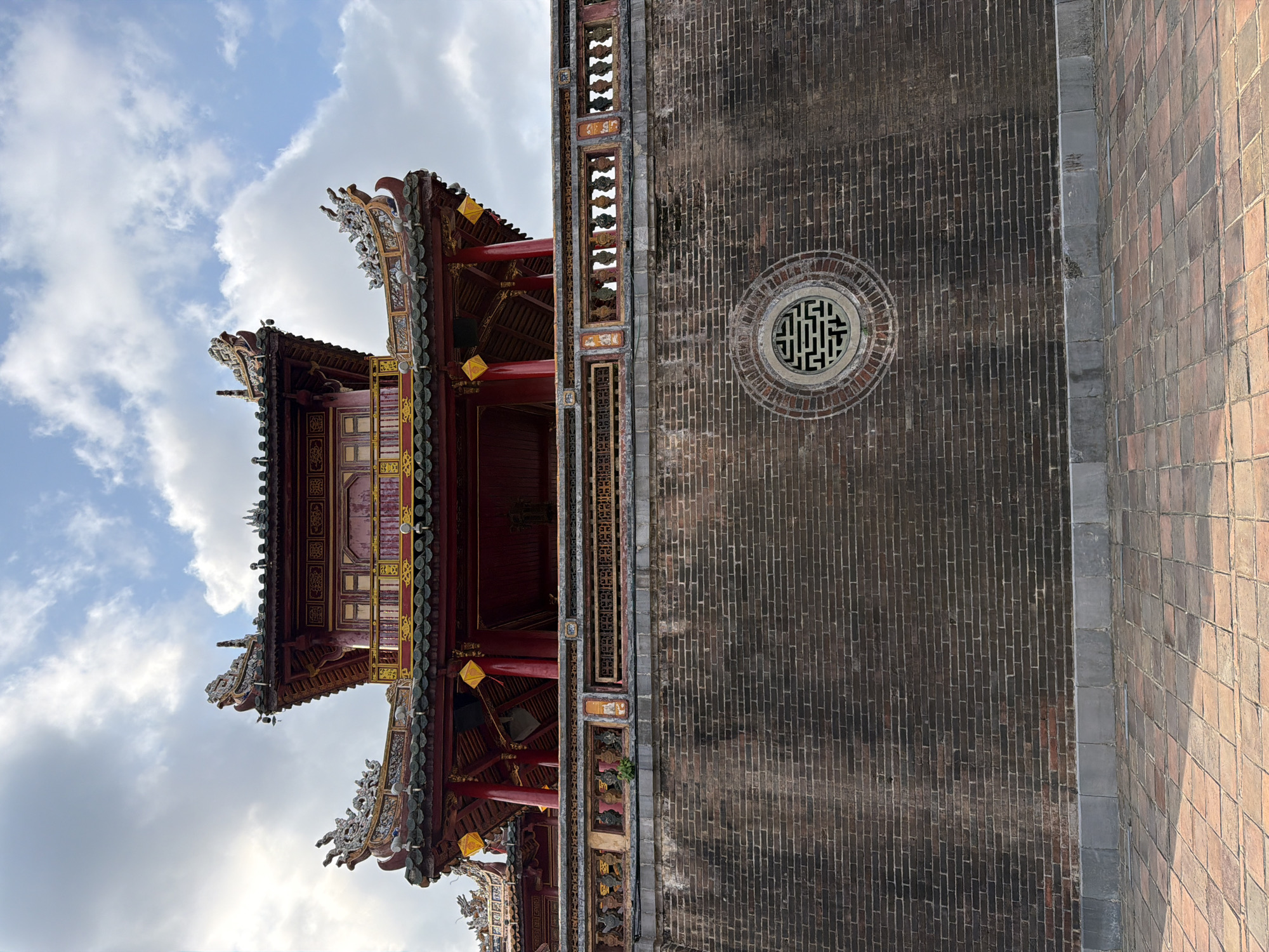 An eye-level, full shot captures the ornate facade of the Imperial City in Phú Xuân Ward, Vietnam, under a partly cloudy sky. The primary subject is a multi-tiered pagoda-style building, predominantly red and gold, with intricately carved dragon motifs adorning its dark grey tiled roofs. The building is elevated, rising above a substantial dark brick wall. This wall, appearing weathered and aged, features a decorative circular vent with a geometric pattern in its center. Below the wall, a paved courtyard of reddish-brown tiles extends towards the viewer, catching the sunlight and creating shadows. The scene is static, with no individuals present. The environment suggests an exterior setting within a historical architectural complex.