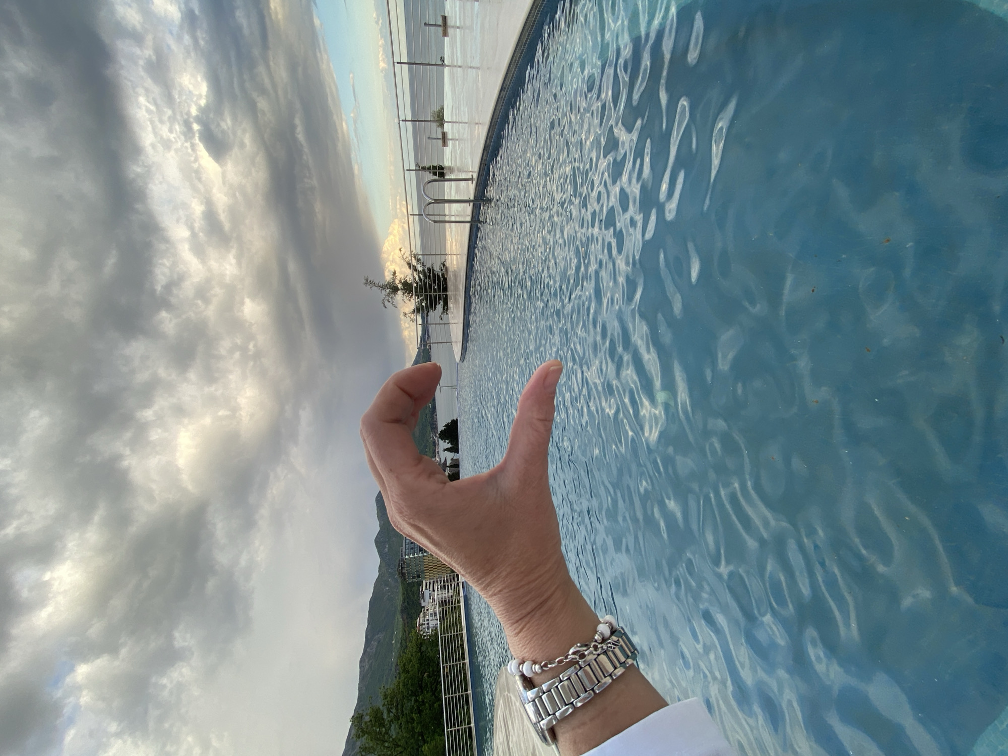 A photograph taken in Boreti, Montenegro, shows a hand with a silver watch and bracelet held up in front of an infinity pool. The pool water is a light blue, reflecting the sky. Beyond the pool, a coastline is visible with some buildings and greenery along the base of a mountain. The sky is cloudy and overcast. The picture is oriented vertically.