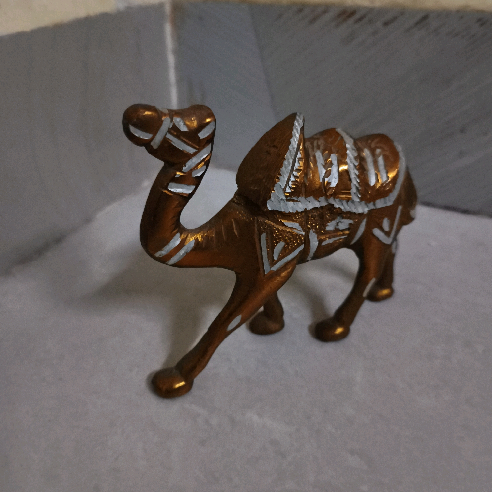 A close-up, eye-level, medium shot captures a small, ornamental camel figurine made of a metallic material with a bronze-like finish. The figurine is detailed with intricate white line patterns adorning its hump, body, and legs, suggesting a decorative, possibly Middle Eastern or North African, motif. The camel is positioned on a flat, light gray surface, which appears to be a tabletop or floor. Behind the figurine, a blurred background shows a textured, light-colored wall or partition, with diagonal lines suggesting either a tiled surface or a shadow pattern. The lighting is moderate, casting a subtle shadow to the right of the figurine, highlighting its three-dimensional form. No people are visible, and no specific location within Mumbai, India, can be identified from the image.