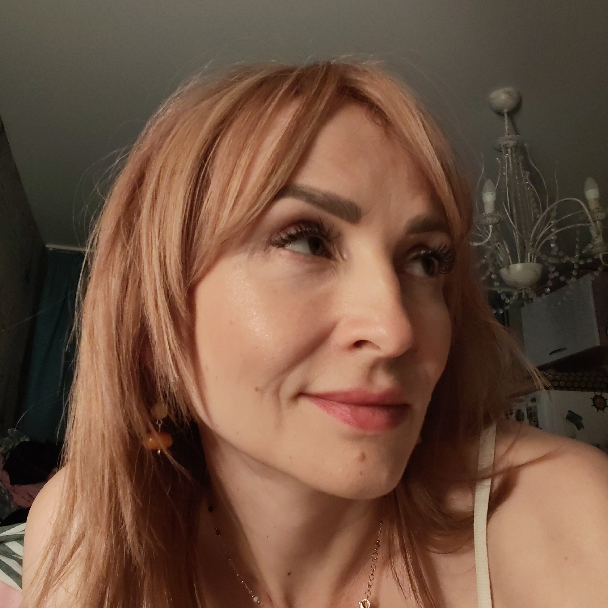 A woman with reddish-blonde hair and bangs is looking off to the side. She is wearing earrings and a necklace. In the background, there is a chandelier and other furniture. The setting appears to be a room indoors. It is not possible to determine if this image was taken in Chaikovsky, Russia.