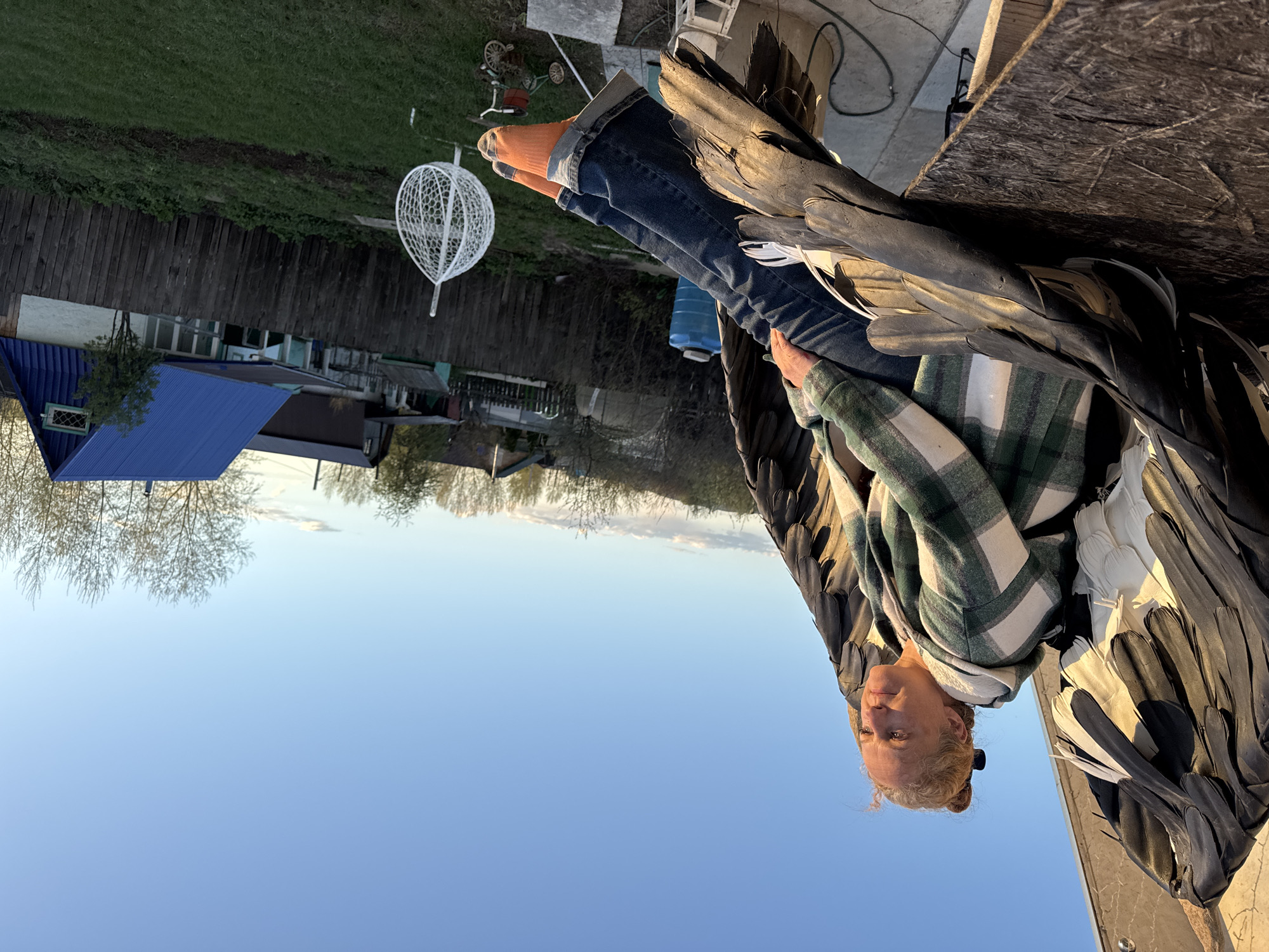 The image depicts a person, possibly female, with blonde hair tied back, wearing a green and white plaid shirt and blue jeans. They are sitting on a wooden structure, with orange socks visible on their feet. The person is adorned with large, stylized wings made of black and white feathers. In the background, there is a yard with a white, cage-like object, a two-wheeled vehicle, and a blue-roofed building. The environment appears to be Chaikovsky, Russia, based on the architecture and vegetation visible. The sky is a clear blue. The photo is taken from a low angle, looking up at the person.