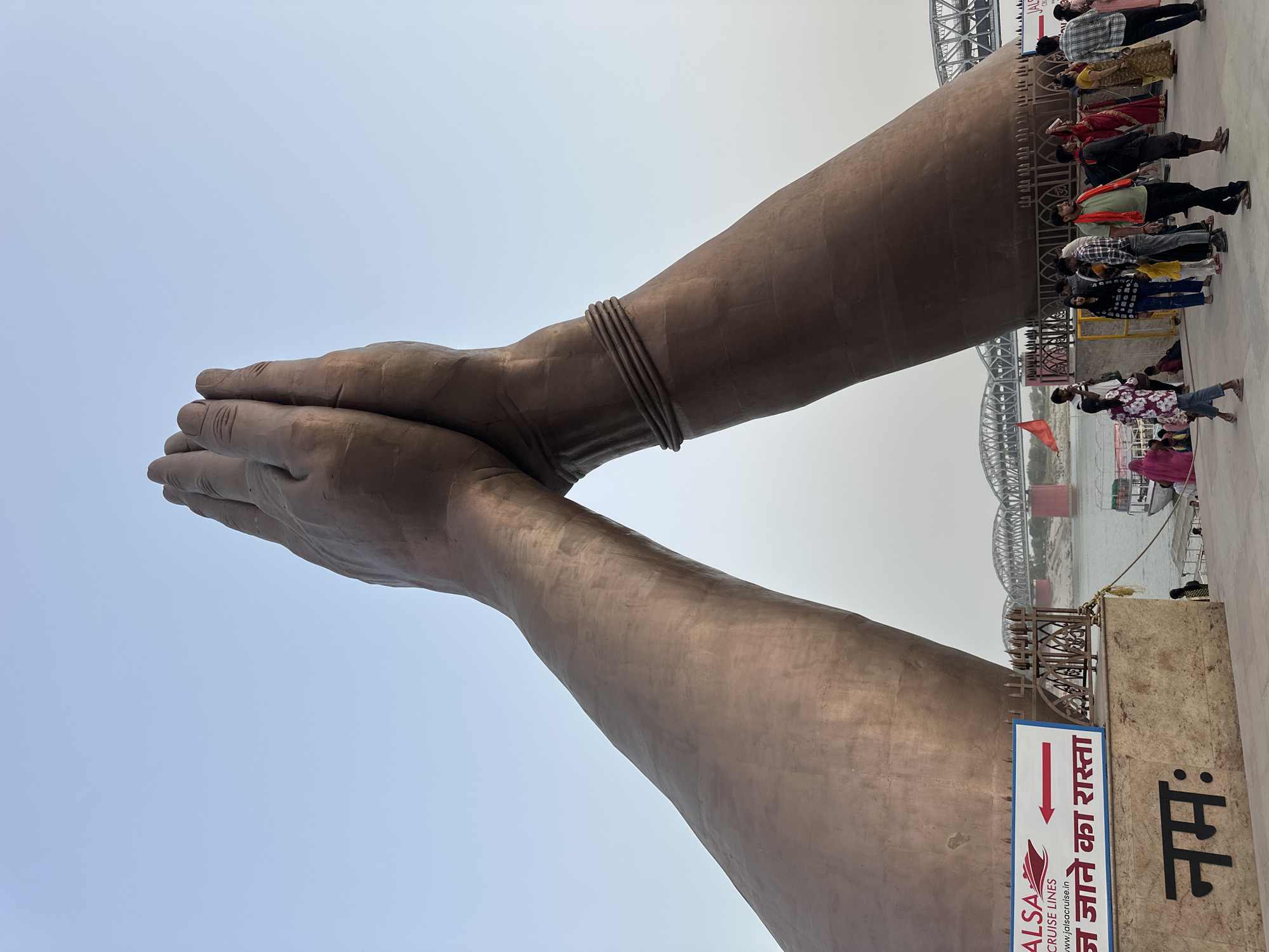 The image depicts a large statue of two hands in a Namaste gesture, likely located in Mughalsarai, India, as inferred from signage. The statue is bronze-colored and detailed with wrinkles and a wristband on one arm. To the right, a metal bridge spans a body of water. People are present on the right side of the statue, some appearing to stand on a viewing platform or structure. A sign, partly visible, contains text in Hindi and English, referencing 'Jalsa Cruise Lines' and directing viewers towards 'जा जाने का रास्ता' (the way to go). The sky is overcast, giving the scene a muted, hazy appearance.