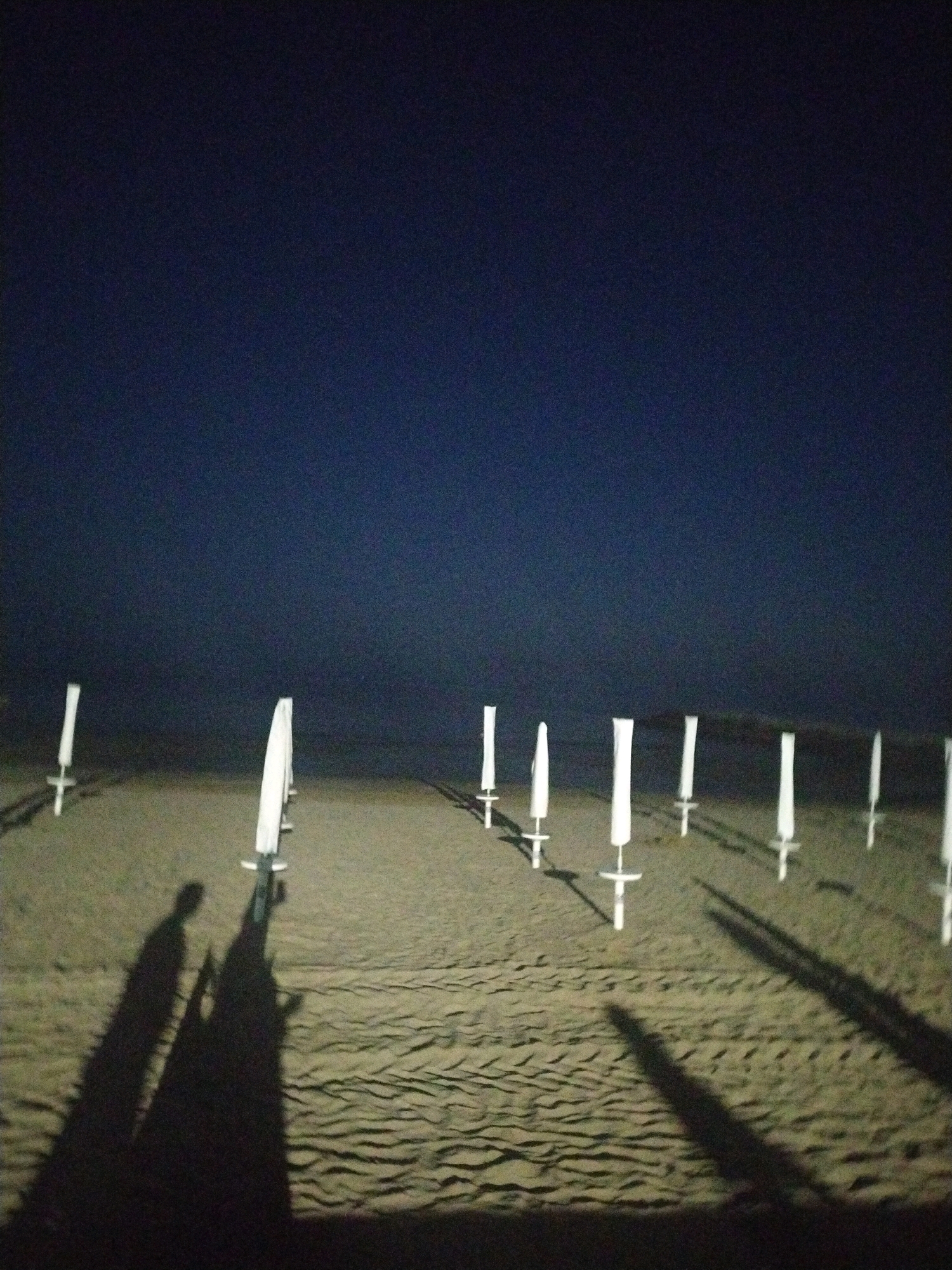 The image depicts a beach scene at night in Caorle, Italy. Several white beach umbrellas, their fabric covers drawn, are planted in the sand, forming a line extending into the distance. The dark sky above is almost entirely devoid of stars. In the foreground, two long shadows are cast across the rippled sand, suggesting the presence of at least two people. The overall impression is one of stillness and quiet, with the minimal lighting creating a stark, almost otherworldly atmosphere.