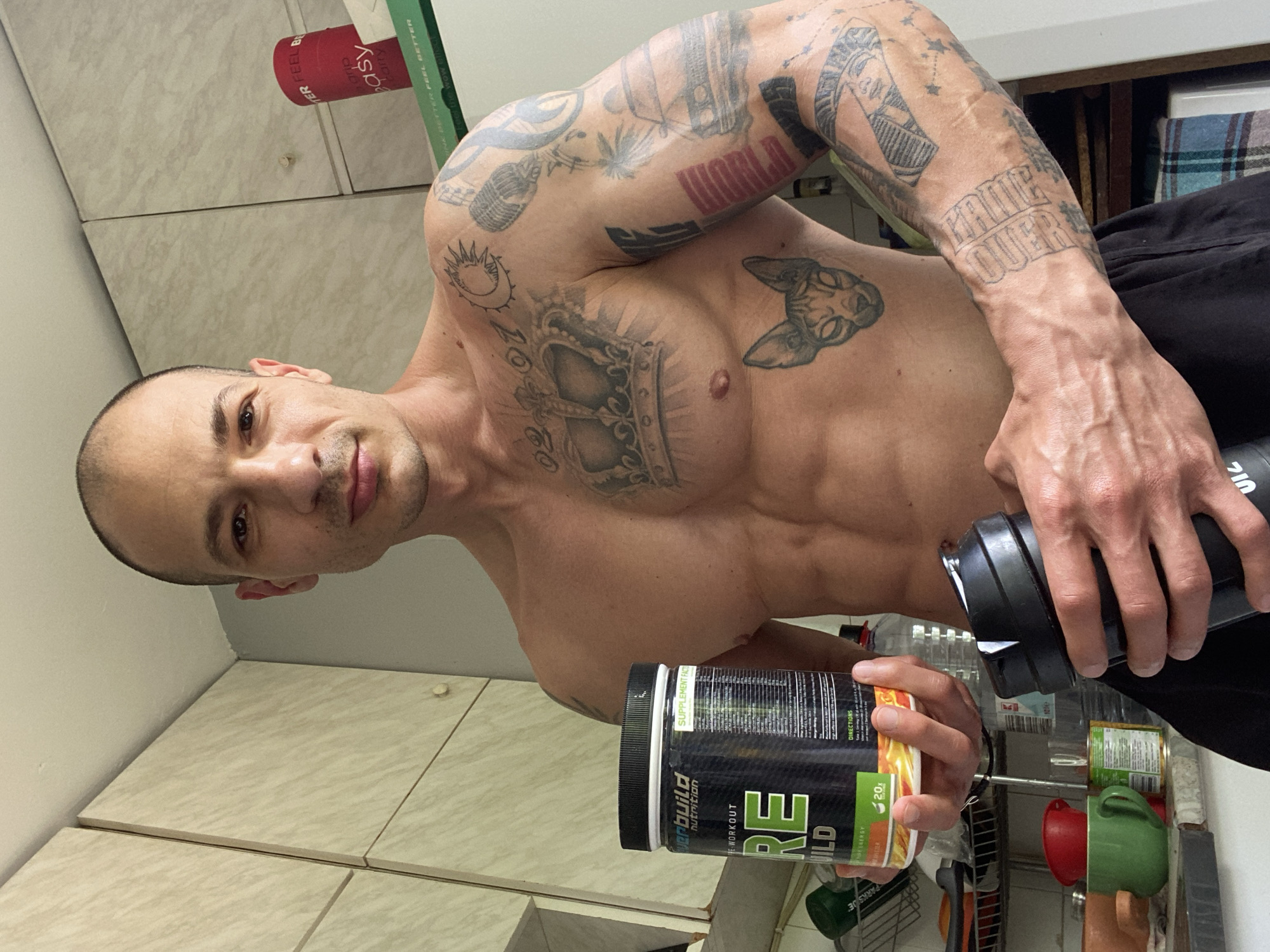 A muscular, bald man with multiple tattoos on his chest and arms is holding a container of "CORE WILD" pre-workout supplement from the brand "verbuild nutrition" in his left hand and a black shaker bottle in his right hand. The man is shirtless and appears to be in a kitchen. Visible in the background are kitchen cabinets, shelves with various objects, and a red "easy carry" grip. The city of Pleven, Bulgaria, is not directly discernible from the image.