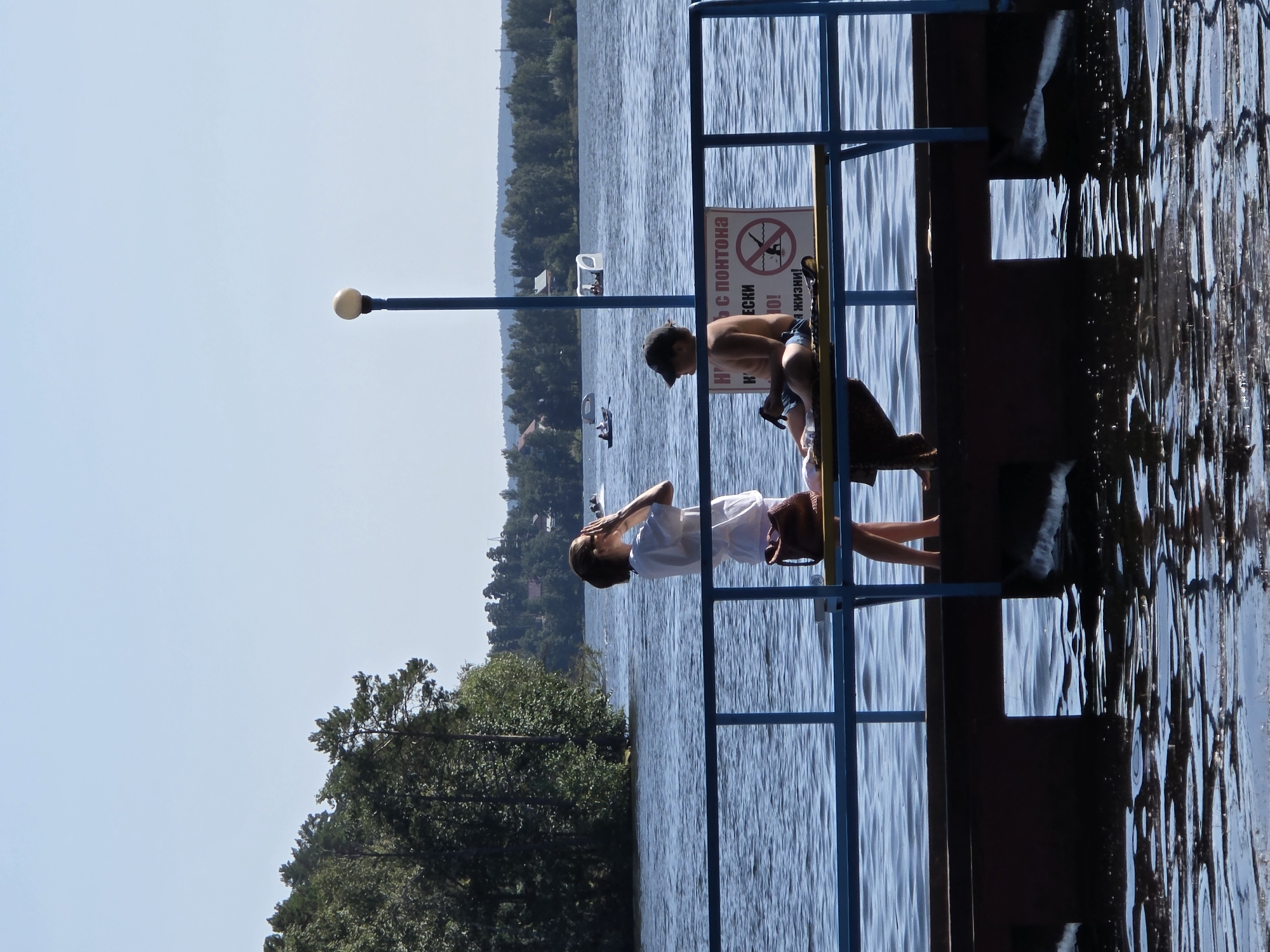 Two people are on a wooden pier over a body of water. A woman in a white t-shirt and shorts stands to the left, looking at her phone with her hands covering her face. A man wearing a black baseball cap and blue shorts is seated to the right, looking to his left. A yellow bench is located on the pier between them. A sign with red and white text and a no-swimming symbol is visible on the pier. The background shows a tree-lined shore with houses and boats on the water under a clear blue sky. The body of water is the Chebarkul Lake in Chebarkul, Russia.