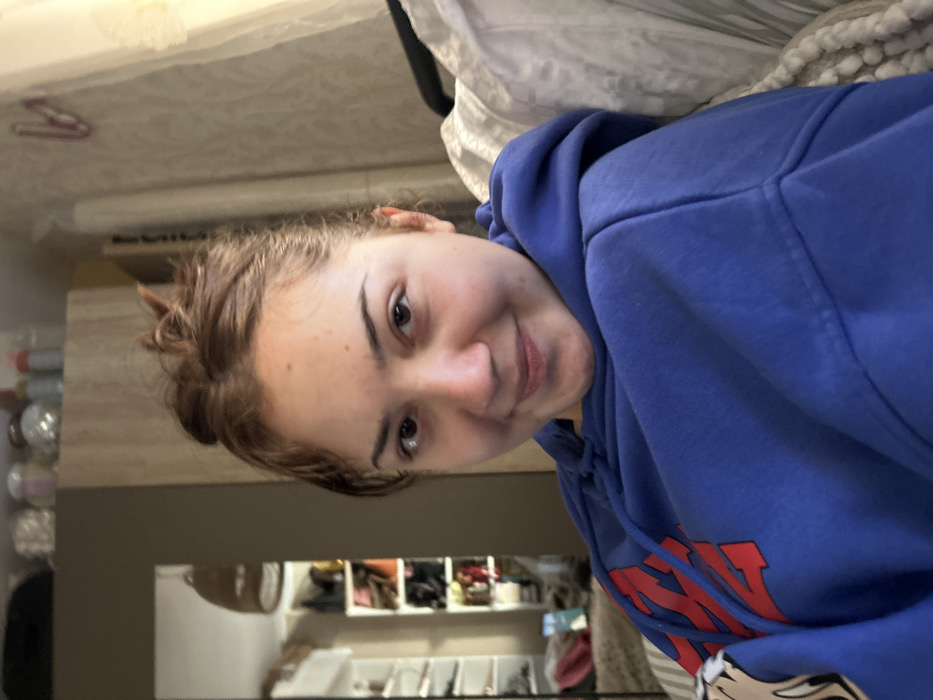 This image portrays a young female positioned centrally indoors, in Schiltigheim, France. The female, appearing to be in her late teens or early twenties, possesses light-toned skin, dark eyes, and light brown hair styled in an untidy bun. She displays a slight smile and is wearing a royal blue hooded sweatshirt with a red graphic visible on the left side of the chest. She appears seated, with her upper body slightly angled towards the viewer.
The background is divided by the subject's presence. To the left, a rectangular mirror reflects a room, showcasing a white open shelving unit or wardrobe containing various pairs of shoes and multiple handbags in different colors. A woven pendant light fixture hangs from the ceiling in the reflection. Directly above the subject's head on the left, a light-colored wooden cabinet or shelf holds several small objects, including glass containers and bottles. To the right of the subject, a wall covered in light-colored, subtly patterned wallpaper is visible. A sheer, light-colored curtain is present on the far right, suggesting a window. Part of a bed or sofa with white and grey patterned textiles is visible in the lower right foreground and within the mirror's reflection. A pale, possibly pink or red, decorative or functional object is affixed to the wall in the upper right quadrant.