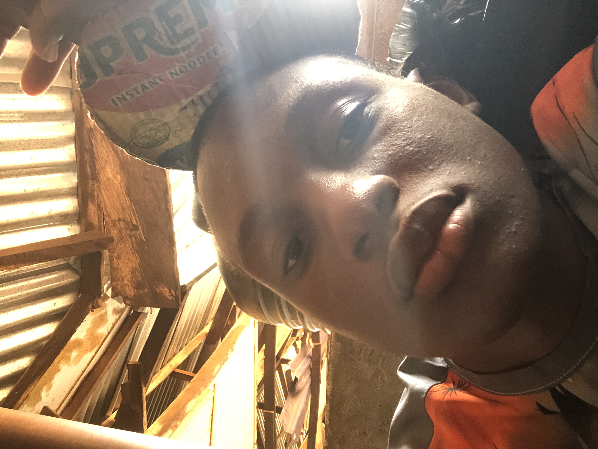 A close-up selfie appears to be taken from a low angle, looking up at a person with dark skin. The person's face fills much of the frame, with the eyes, nose, and lips clearly visible. Above their head is a damaged packet of instant noodles. The background shows a roughly constructed structure, possibly a roof, made of corrugated metal sheeting and wooden supports. The location is implied to be Ukwu-Abwa, Nigeria.