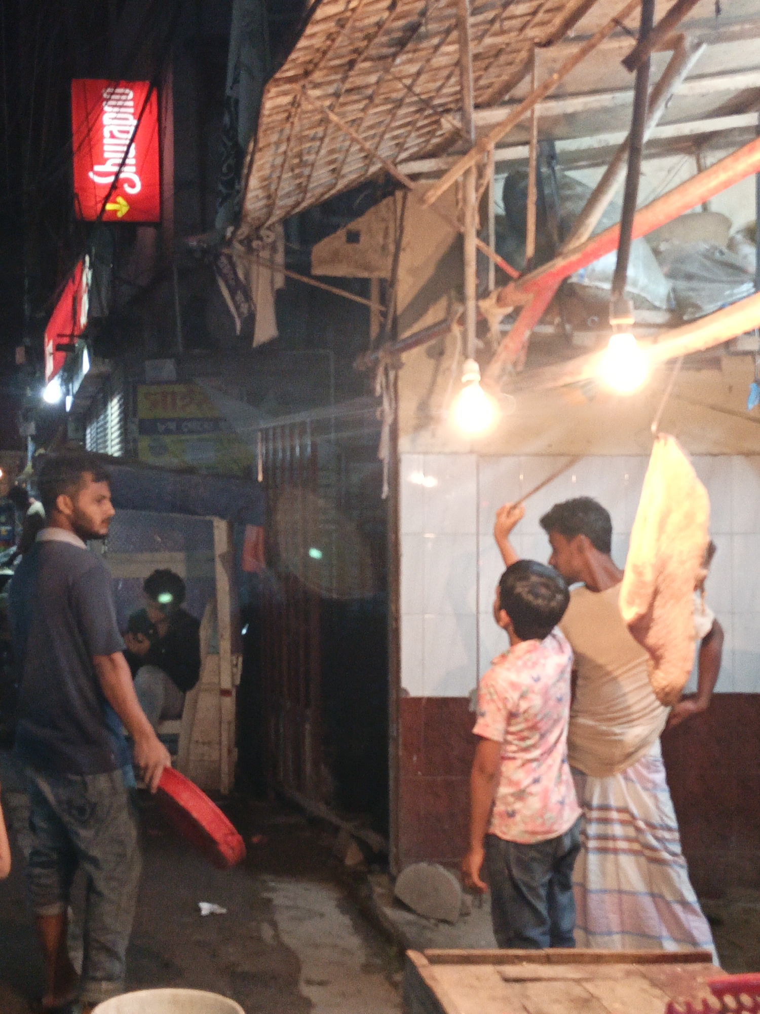 This nighttime image depicts a bustling street or market scene, likely in an urban area of South Asia, illuminated by glowing signs and bare electric bulbs hanging from wires. The setting features temporary or aged structures, characterized by exposed wooden beams and makeshift canopies overhead.
Several men are present and engaged in various activities. On the left, a man with a beard, dressed in a dark blue polo shirt and jeans, stands looking towards the right, holding a red oval-shaped object, possibly a tray or lid. Behind him, inside a dimly lit structure, another person is seated, their face subtly illuminated by a green glow, possibly from a screen.
On the right side of the frame, two individuals are actively involved. An adult male, wearing a light-colored shirt and a traditional checked lungi, holds a long stick with his arm raised, seemingly interacting with something overhead. In front of him, a younger person in a pink floral patterned shirt and jeans looks upwards in the same direction. A light-colored object, possibly a textile or piece of food, hangs near them. Another figure is partially visible behind the man in the lungi.
The ground appears damp and somewhat cluttered, with a white bucket in the lower foreground. The time of day is clearly night, as indicated by the darkness and reliance on artificial lighting. A prominent red neon sign on the left displays white Bengali text that reads "Shurabhi" (শুভ্র), accompanied by a yellow arrow pointing right. Another yellow sign in the background shows partial Bengali script, including the word "Market" (মার্কেট). The scene conveys a sense of daily life and commerce in a lively, informal environment.