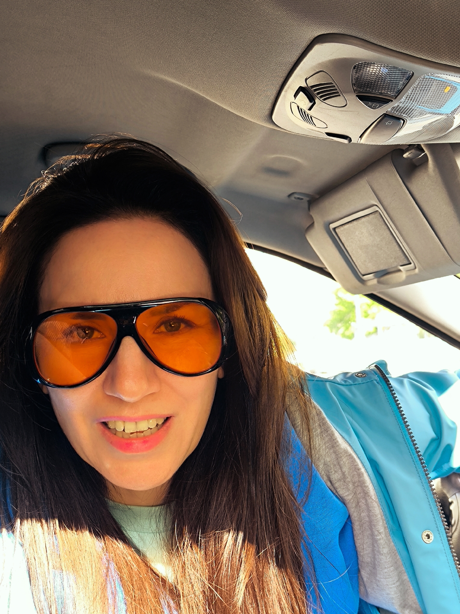 A woman is taking a selfie from inside a car. She is wearing orange-tinted aviator sunglasses, a light blue jacket over a gray sweatshirt, and pink lipstick. The car's interior ceiling, sun visor, and interior lights are visible. The outdoor background is blurry but suggests a daytime setting, possibly in Riga, Latvia.