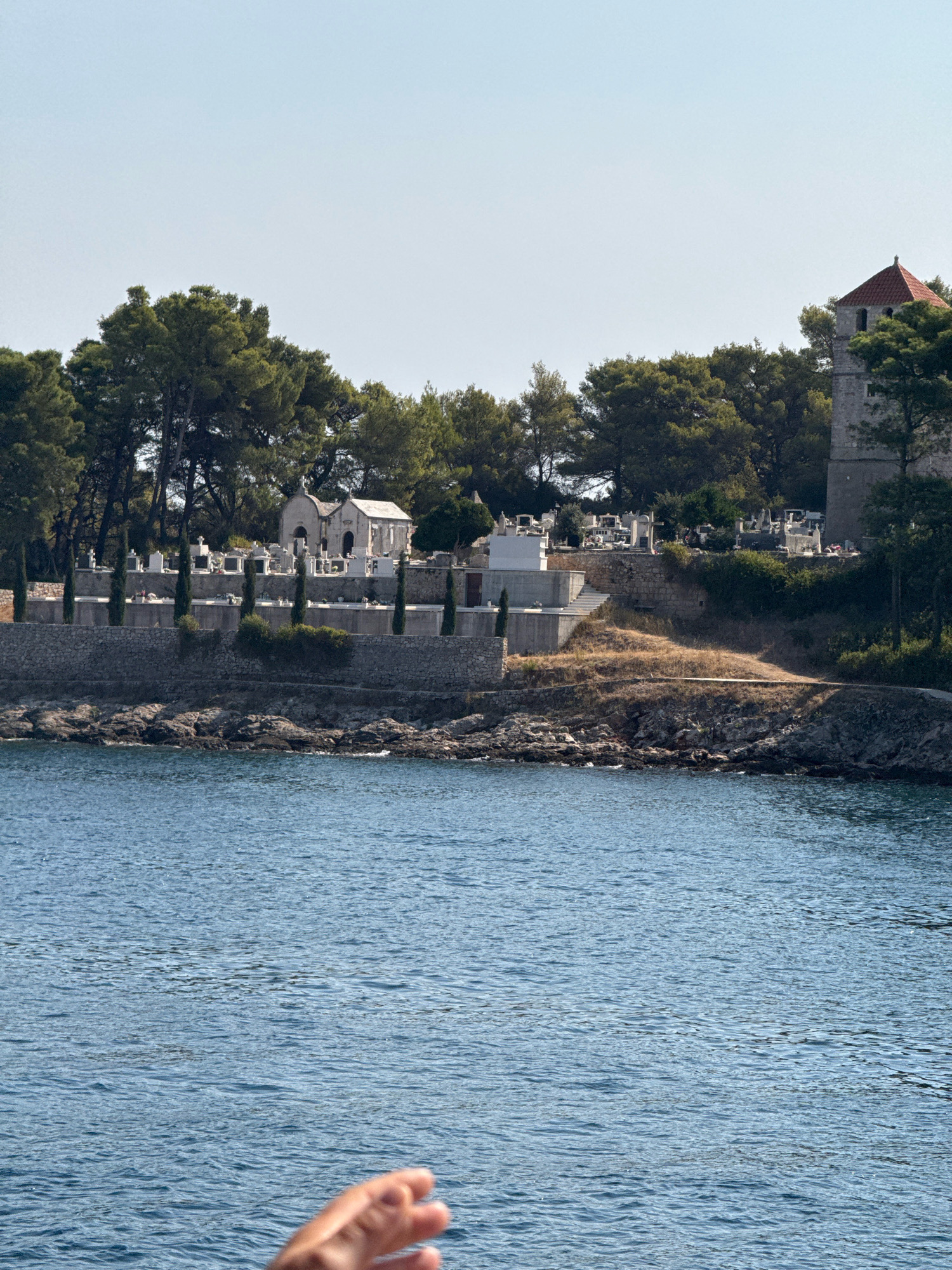 This image captures a view of a cemetery and a bell tower overlooking the sea in Jelsa, Croatia. The cemetery is situated on a hillside, with numerous gravestones and a small chapel visible. Cypress trees are planted throughout the cemetery, and a stone wall borders the property. The bell tower, with its red-tiled roof, stands tall among the trees. The sea in the foreground is a deep blue, with small waves lapping against the rocky shore. A hand is visible in the lower right corner of the image, suggesting the photo was taken from a boat or a similar vantage point. The sky is clear and blue, indicating a sunny day.