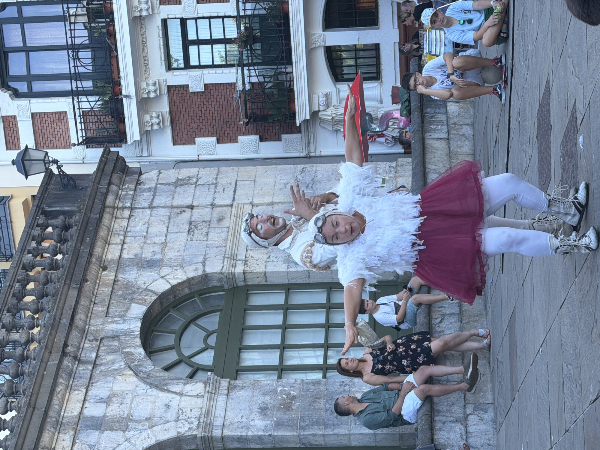 In Lekeitio, Spain, in front of a stone building with arched windows, two performers are engaged in a street performance. The performer in the foreground is wearing a white feathery costume, a burgundy tutu, white leggings, and combat boots. She is in the process of a dramatic pose, with her arms outstretched to the sides and her head tilted back. Behind her, another performer, dressed in a white outfit with goggles and white face paint, gestures animatedly with his hands. Several spectators are seated on a stone ledge in the background, watching the performance. To the right, a young boy wearing glasses sits on a ball, looking bored, while another boy in a blue shirt and hat watches the performance. The scene is set in a public square with cobblestone paving.