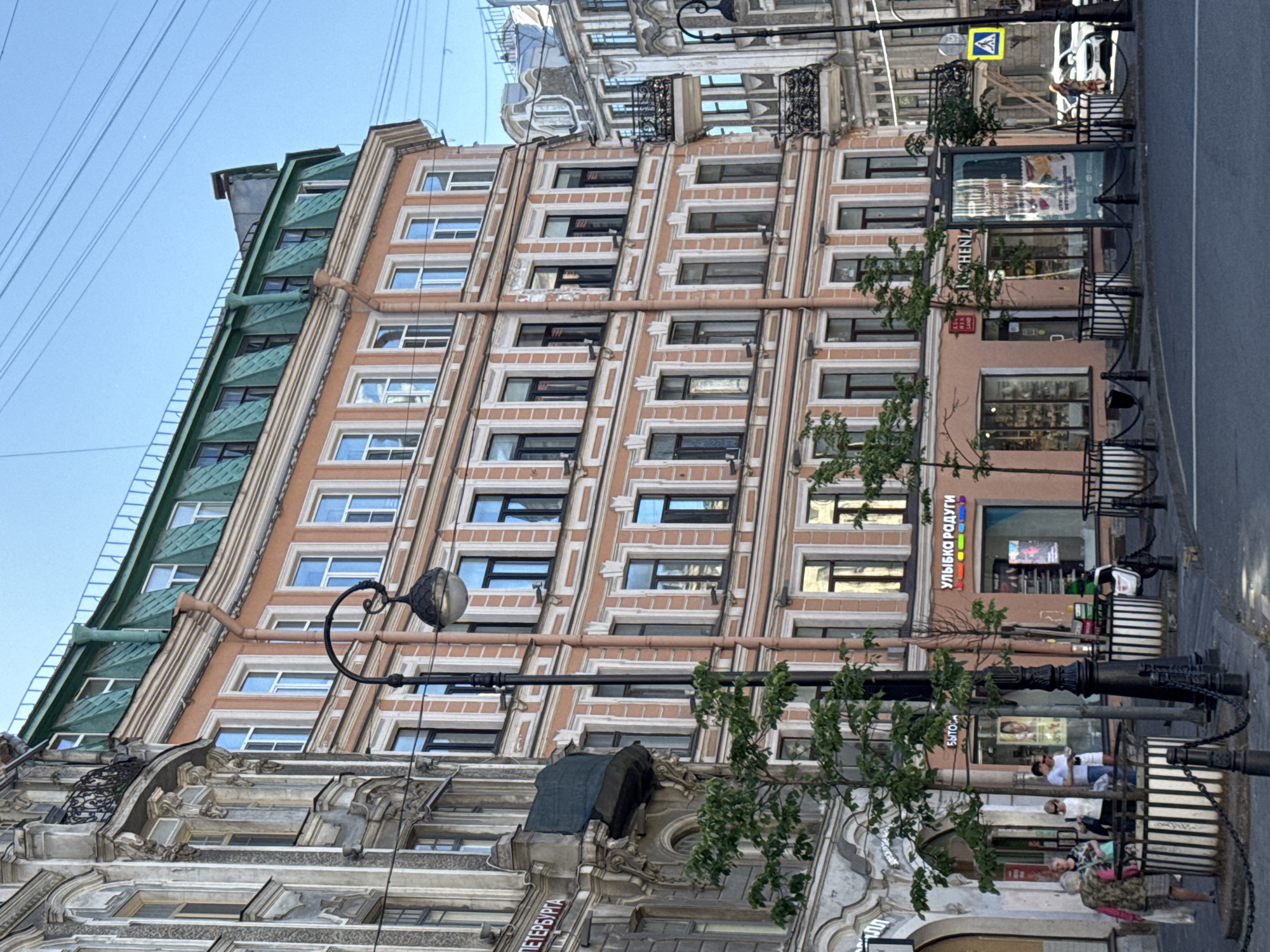 This is an urban street scene captured during daylight hours under a clear blue sky. The image focuses on a row of multi-story historical buildings in Saint Petersburg, Russia, indicated by the visible text "ПЕТЕРБУРГА" on the left-most building and "УЛЫБКА РАДУГИ" (Ulybka Radugi - a Russian chain store) on the ground floor of the central building.
In the center, a prominent six-story building with a light salmon-colored facade and white architectural trim occupies the midground. It features numerous rectangular windows with dark frames and a distinctive green mansard roof with dormer windows. Vertical drainage pipes run down its facade. The ground floor houses commercial establishments with large display windows, including a store signed "УЛЫБКА РАДУГИ" and another partially visible as "CHENZLAND."
To the left, a more ornate, grayish multi-story building with classical architectural details, including decorative moldings and a wrought-iron balcony, is partially visible. To the right, another ornate gray building with balconies and intricate ironwork is visible in the background.
The street in the foreground features asphalt pavement with white lane markings. A paved sidewalk lines the buildings, furnished with black, ornate streetlights, one centrally placed with a spherical lamp. Striped black-and-white planters containing small green trees are chained off with black bollards. A large, vertical outdoor advertising display stands on the right side of the street, partially obscuring a white SUV. A blue pedestrian crossing sign with a yellow triangle is visible on the far right. Numerous dark overhead utility wires crisscross the bright blue sky.
Several people are present on the left sidewalk: a group of approximately five adults is conversing near a planter, including a woman in a patterned top, a man in a white t-shirt, and another woman holding a light-colored bag. Another person in light clothing is partially visible walking on the right sidewalk, behind the white car.