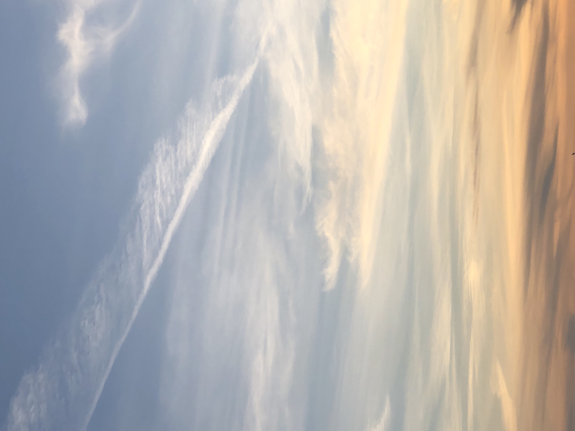 The image depicts a sky with a gradient of colors, transitioning from a light blue to a mix of yellow and orange hues on the right side. The sky contains wispy clouds scattered throughout, and a prominent contrail is visible in the left side of the image. The photo was taken in Krasnoyarsk, Russia.