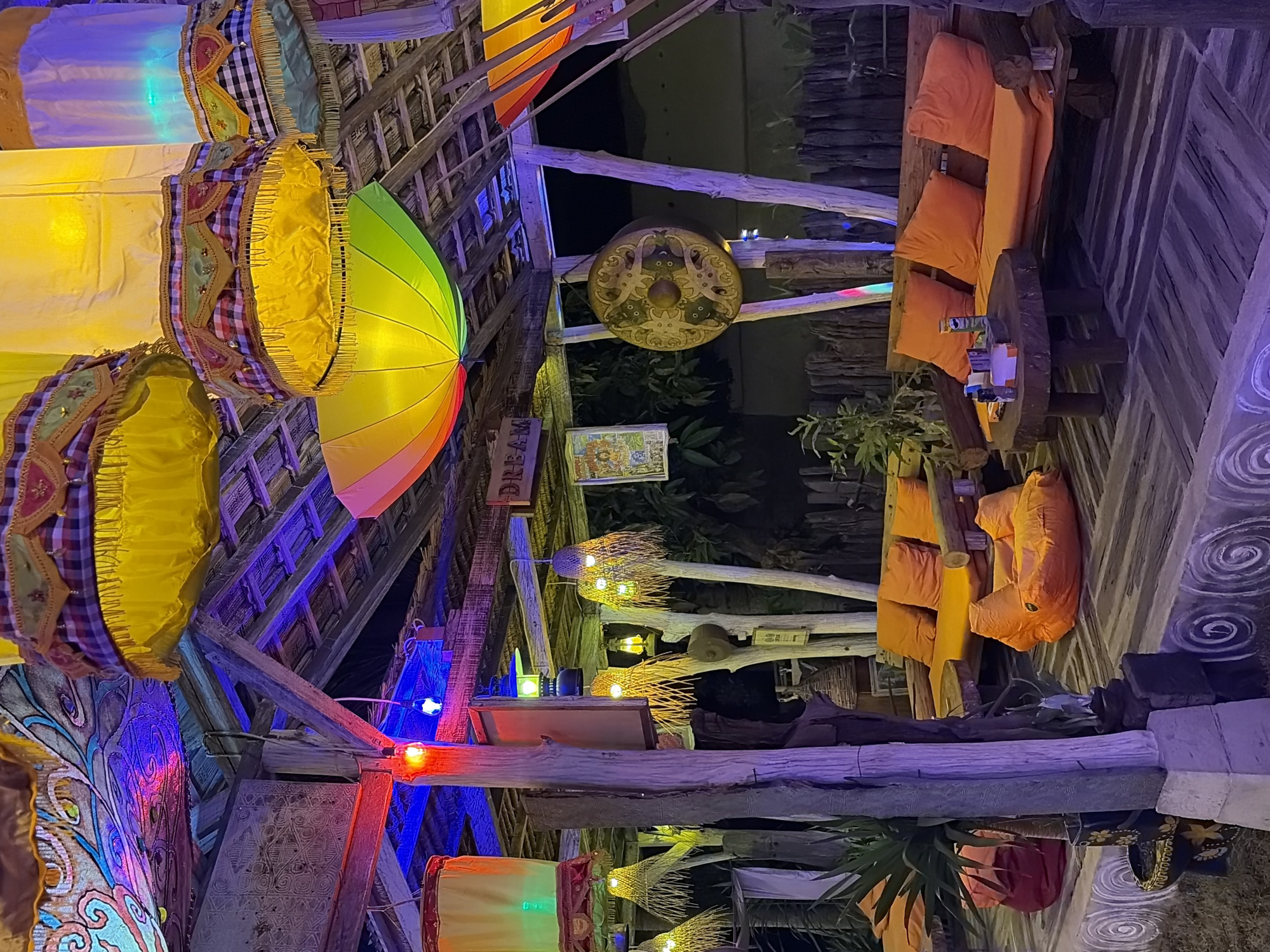 This is a medium shot, eye-level photograph of an outdoor seating area with rustic wooden furniture and vibrant lighting in Banjar, Indonesia. The scene is illuminated by a mix of colorful lights, including red, blue, yellow, and purple, which cast a vibrant glow on the wooden structure and furnishings.
In the foreground, there are two large wooden benches with bright orange cushions, accompanied by matching orange beanbag chairs. A low, round wooden table sits between the benches, with a few bottles and containers on its surface. The floor is made of wooden planks, with a patterned concrete border.
Above the seating area, a thatched roof is supported by wooden beams. Hanging from the roof are several large, decorative lanterns. Some are cylindrical and yellow, adorned with patterned fabric and fringe, while one is a colorful, umbrella-like structure with rainbow stripes. Other smaller, woven lanterns hang lower, casting a warm yellow light.
The background reveals more of the rustic wooden structure and a dark, natural environment, suggesting a tropical outdoor setting. A large, circular gong with an intricate design is visible hanging on a wooden post. The overall impression is one of a lively and decorative outdoor entertainment or dining space.