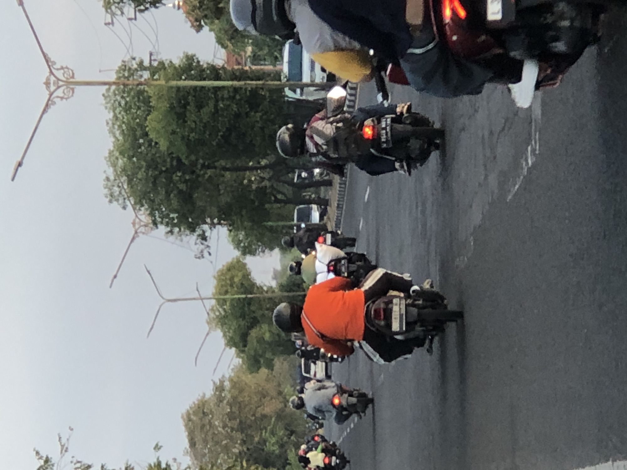 The image depicts a street scene in Siring, Indonesia, with a convoy of motorcycles navigating a paved road. The sky is overcast, suggesting either early morning or late afternoon. Dominating the foreground is a motorcycle rider seen from behind, wearing an orange t-shirt and black pants, astride a dark-colored motorcycle. Their right leg is extended, and a silver crossbody bag is slung over their shoulder. To the left and slightly ahead, another rider in a grey shirt and black pants is also viewed from behind, their motorcycle's red taillight illuminated. Further ahead, a rider in a grey and maroon jacket with a backpack is observed from the side. A motorcycle in the far right foreground, partly obscured, shows a rider wearing a blue jacket and a grey helmet. Several other motorcycles and riders are visible in the middle ground and background, moving in the same direction. White lane markings are visible on the asphalt road. Trees line the sides of the road, with some streetlights with decorative metalwork interspersed among them. In the background, several white vehicles and more trees are present. The overall impression is of moderate traffic flow on a city street.