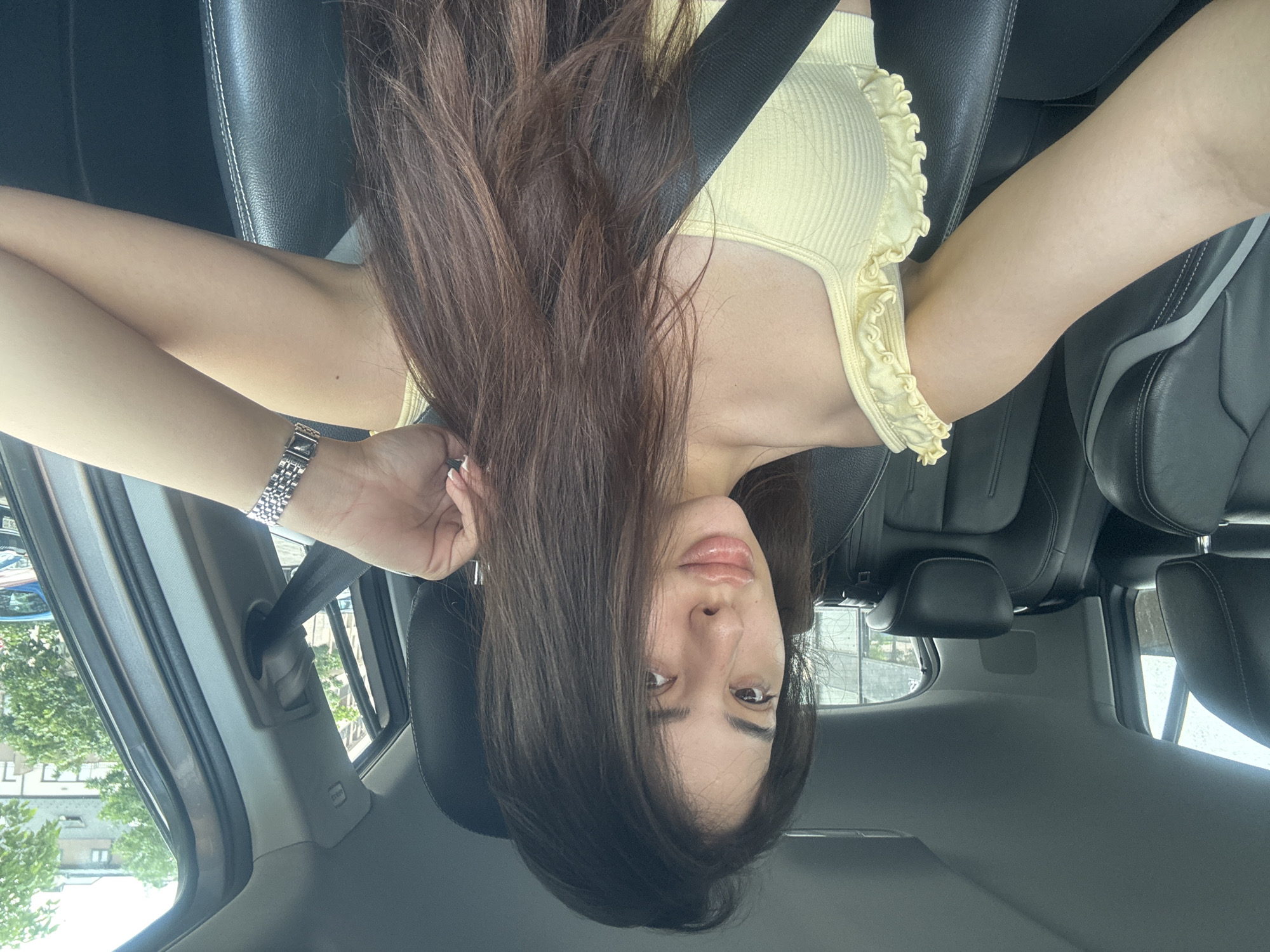 A young woman is prominently featured in a close-up photograph taken inside a car, with the image rotated 90 degrees clockwise from a standard portrait orientation. She is looking upwards towards the camera, suggesting she is reclining or the photo was taken from above. Her long, dark brown hair cascades downwards across a significant portion of the frame, framing her face. She has a calm, neutral expression, looking directly at the viewer with slightly parted, glossy lips.
She is wearing a light yellow, ribbed tank top with distinctive ruffled trim along the straps and bottom hem. A silver-toned watch with a segmented metal band adorns her left wrist. The setting is clearly a car interior, visible through the black leather seats with white stitching, headrests, and a seatbelt stretched across her chest. Through the car windows, an outdoor scene reveals buildings and lush green trees, characteristic of an urban environment like Plovdiv, Bulgaria. The bright, natural light indicates it is daytime. The image captures a posed moment, likely a selfie. No discernible text is visible in the frame.