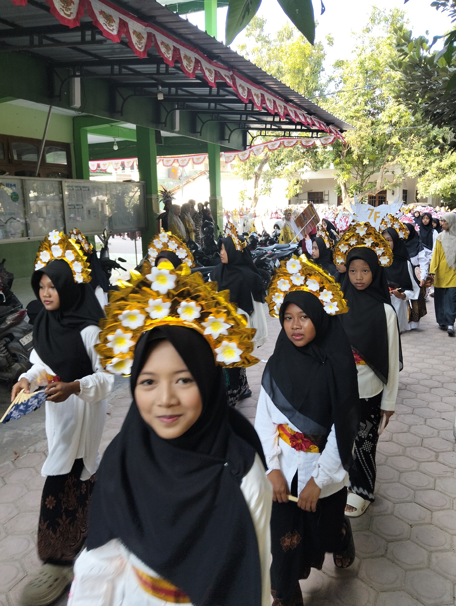 In Kranji, Indonesia, a group of young women are participating in a parade. They are dressed in traditional attire consisting of white tops, black hijabs, and patterned skirts. Each woman wears an elaborate headdress adorned with gold leaves and white flowers. Some women are holding fans, suggesting a celebratory or cultural event. They are walking on a paved walkway lined with trees. In the background, a building with red and white banners and green trim is visible, along with parked motorcycles. The scene appears to be set during the daytime with bright sunlight casting shadows.