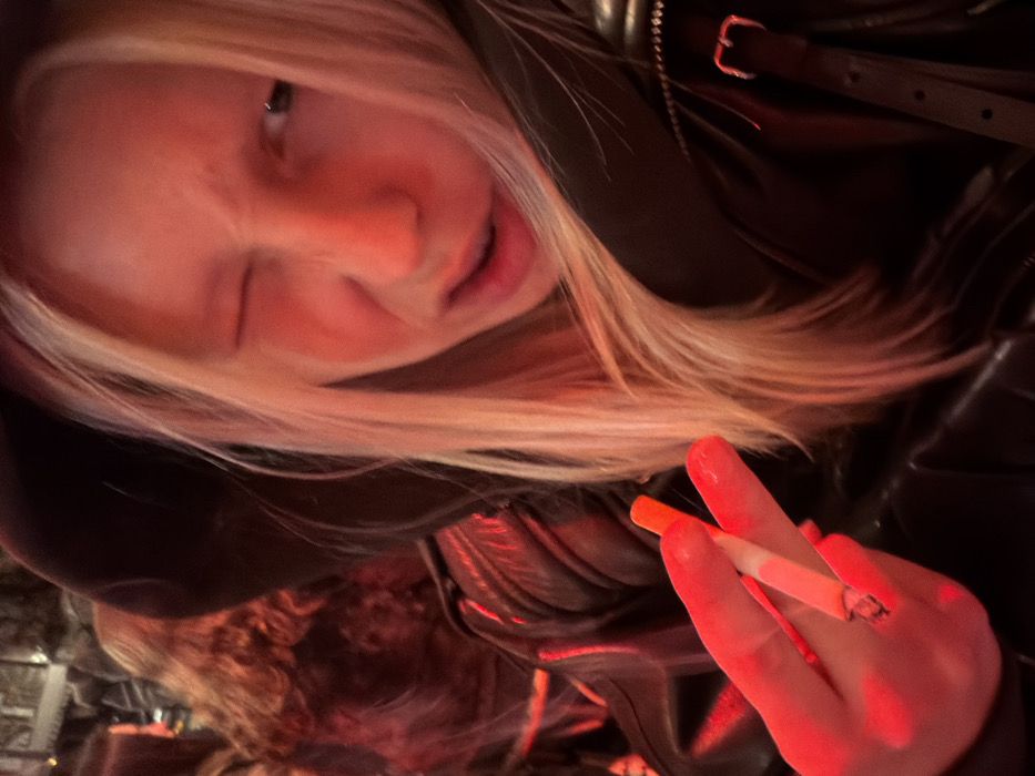 A woman with blonde hair, wearing a dark leather jacket, holds a lit cigarette between her fingers. Her face is close to the camera, with a slight wink and smirk. The lighting is red, highlighting her skin and clothing. A buckle on the jacket strap is visible. There's a glimpse of someone with curly hair in the lower left corner.