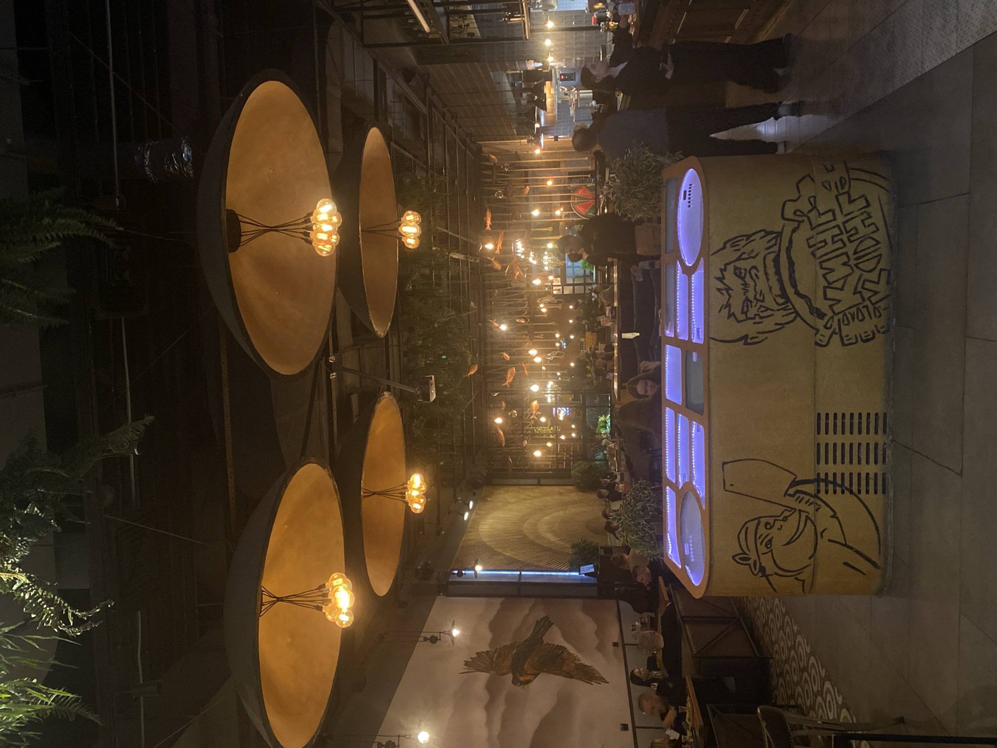 The image captures the interior of the "Estory Bar" located in the city of Yekaterinburg, Russia. The interior features oversized, circular light fixtures with visible filament bulbs, arranged in a vertical row. The restaurant has a contemporary design with exposed metal beams and a high ceiling. A light-colored cabinet with blue lights on its front is decorated with cartoon-style graphics of human figures. The walls are adorned with artwork, including a large painting of a bird in flight. Numerous patrons are seated at tables, suggesting a busy environment. Overhead, decorative lighting elements are suspended from the ceiling. The color palette is dominated by warm tones from the lighting and fixtures, contrasted with darker shades from the architectural elements.