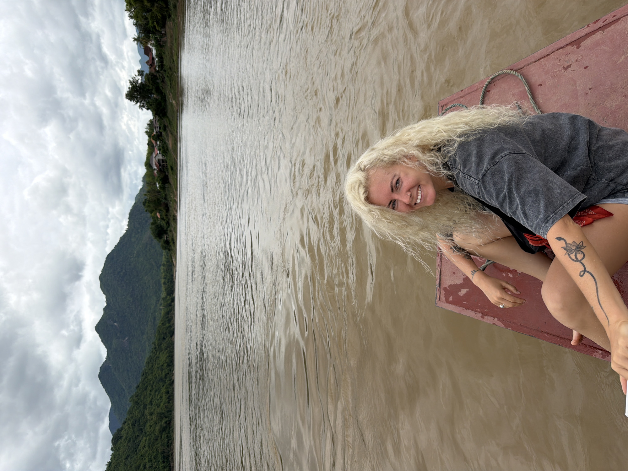 In Houaymat, Laos, a blonde woman with curly hair and a nose ring smiles as she sits on the edge of a flat-bottomed boat. The boat, which appears to be made of wood with a red painted surface, is on a wide, muddy brown river. The woman is wearing a grey t-shirt and denim shorts, and has tattoos on her left arm. Her hands rest on her lap, and she is looking directly at the camera. In the background, lush green mountains rise up, covered in dense vegetation. A small village with houses and buildings is visible along the riverbank, nestled between the mountains and the water. The sky is overcast with grey clouds, suggesting a humid climate.