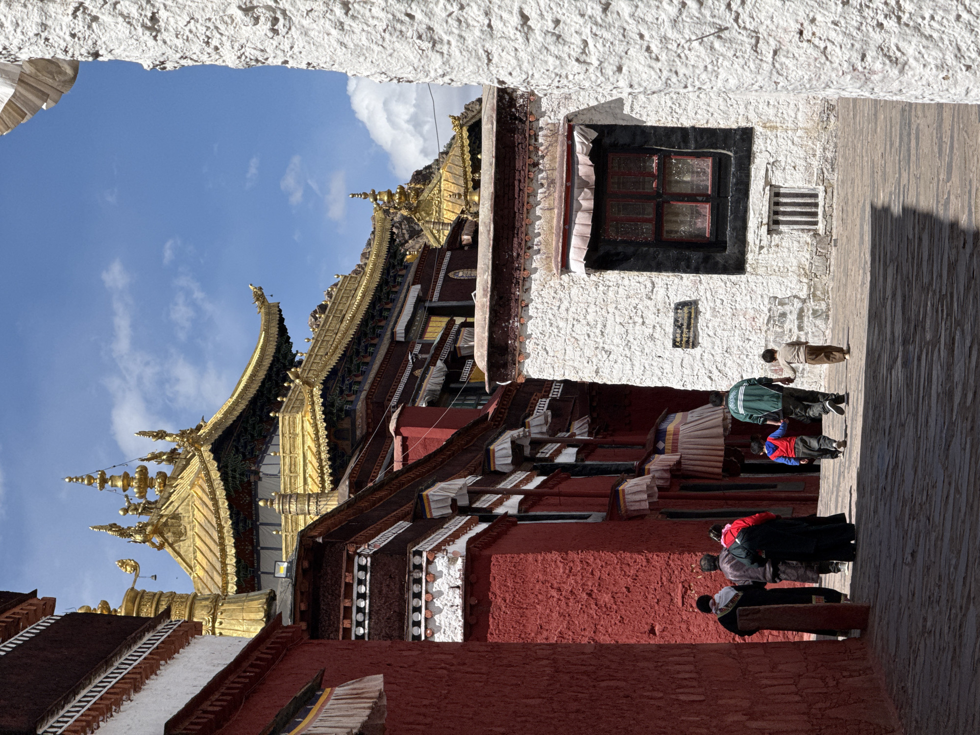 The image captures a street scene in 城西街道, China, featuring a traditional architectural structure with golden roofs and red and white walls. The building appears to be a temple or historical site. A group of people is seen walking along the street. The photo is taken through an archway, creating a framed view of the scene. The sky is visible in the background.