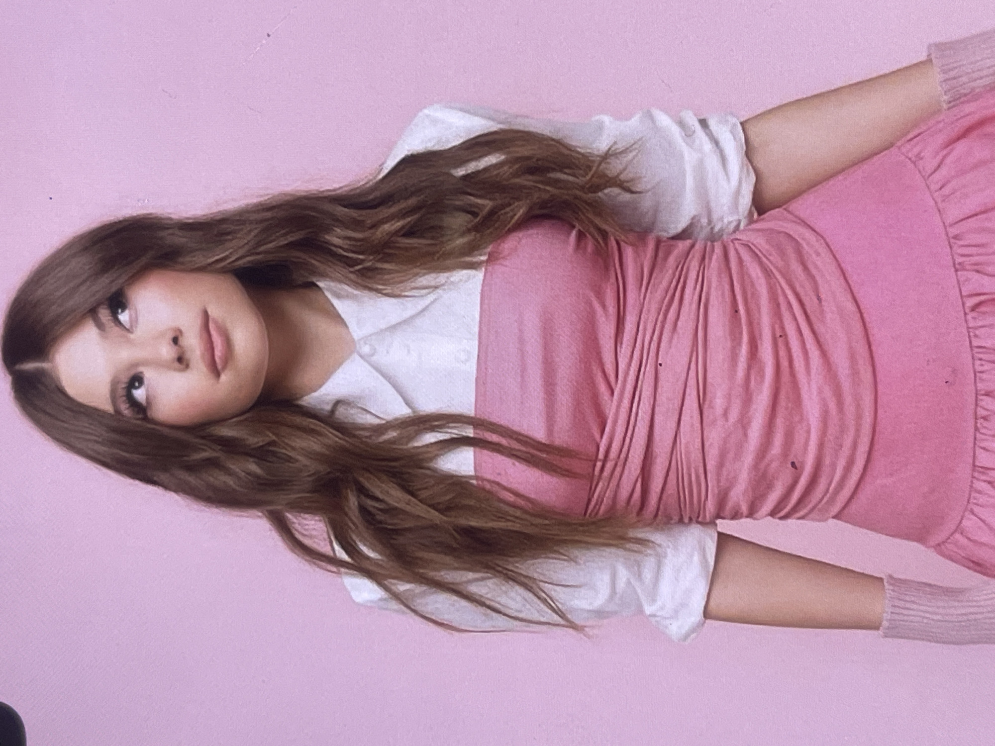 A young female is depicted in what appears to be a studio portrait, lying down against a plain, solid light pink background. Her head is angled towards the top-left of the frame, and she gazes upwards and slightly to her left with a gentle, contemplative expression. She has long, wavy, medium-brown hair that spreads out around her head and over her shoulders.
She is dressed in a layered outfit: a white collared shirt worn beneath a pink garment that resembles a strapless top or dress, featuring gathered fabric on the bodice and a ruffled hemline visible towards the bottom. She also wears light pink, knitted arm warmers or fingerless gloves on both forearms.
The setting is minimalist, consisting only of the uniform pink backdrop, suggesting an indoor studio environment or a specially painted wall. No other objects or specific location cues are visible within the image. The lighting is soft and even. No text is visible. There are no visual elements within the image to indicate time of day, weather, or a specific geographic location such as Poltava, Ukraine.