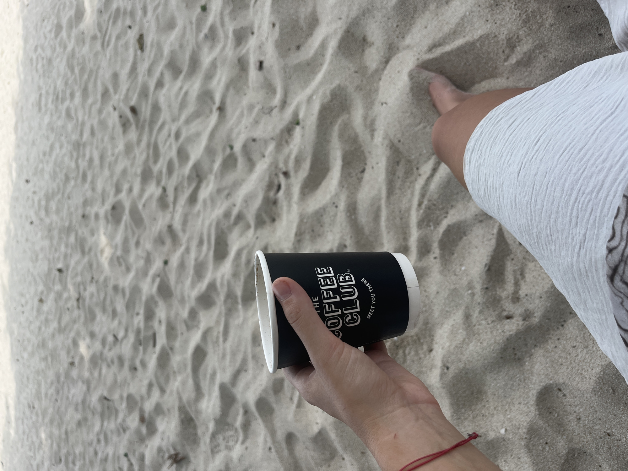 A person's left hand holds a black paper coffee cup with white text that reads "THE COFFEE CLUB MEET YOU THERE." The hand has a thin red string bracelet on the wrist. The person is sitting on a sandy beach, with their legs visible, covered by a white textured skirt or fabric. The sand is light brown and has visible ripples and small debris. The scene is outdoors, suggesting a beach location, likely in Baan Chaweng, Thailand.