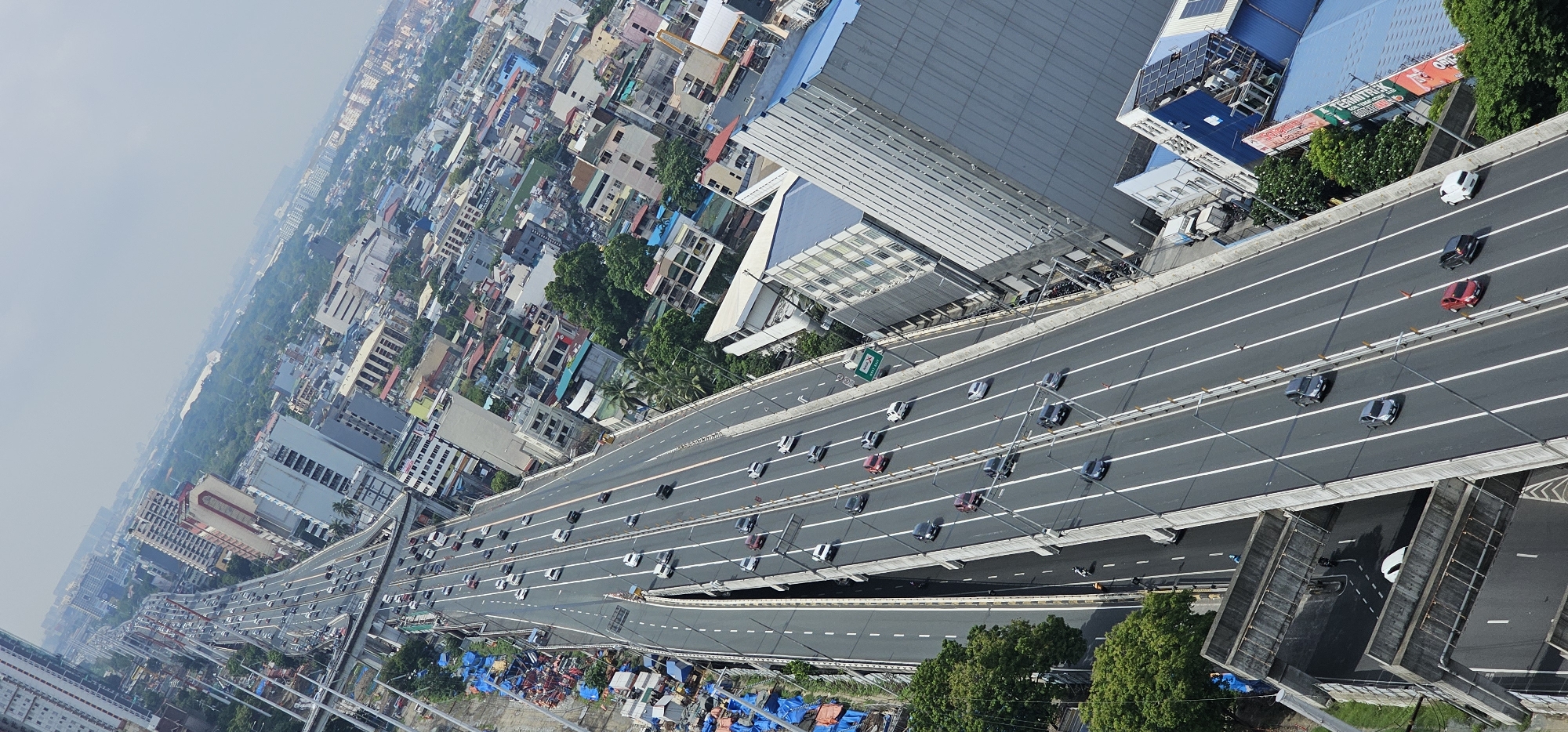 An elevated highway, likely the Skyway System in Makati, Philippines, stretches diagonally across the frame from the lower left to the upper right. The highway is busy with traffic, featuring multiple lanes of cars and trucks. The roadway is a dark asphalt surface marked with white lines.
On the left side of the highway, below the elevated structure, is an area with what appears to be informal settlements or storage, covered with blue tarps and various materials. Adjacent to this area is a vibrant green canopy of trees.
To the right of the highway, the cityscape unfolds. A mix of modern and older buildings, varying in height and architectural style, are densely packed. Some buildings are taller, with multiple stories and glass windows, while others are smaller and more traditional. One prominent building on the right is a large, modern structure with a distinctive blue roof and solar panels, possibly a commercial or industrial building. A large red and white billboard is visible on the side of this building.
The sky is overcast, with a hazy, bright atmosphere suggesting a sunny or humid day. The overall impression is one of a bustling urban environment with extensive infrastructure and high population density.