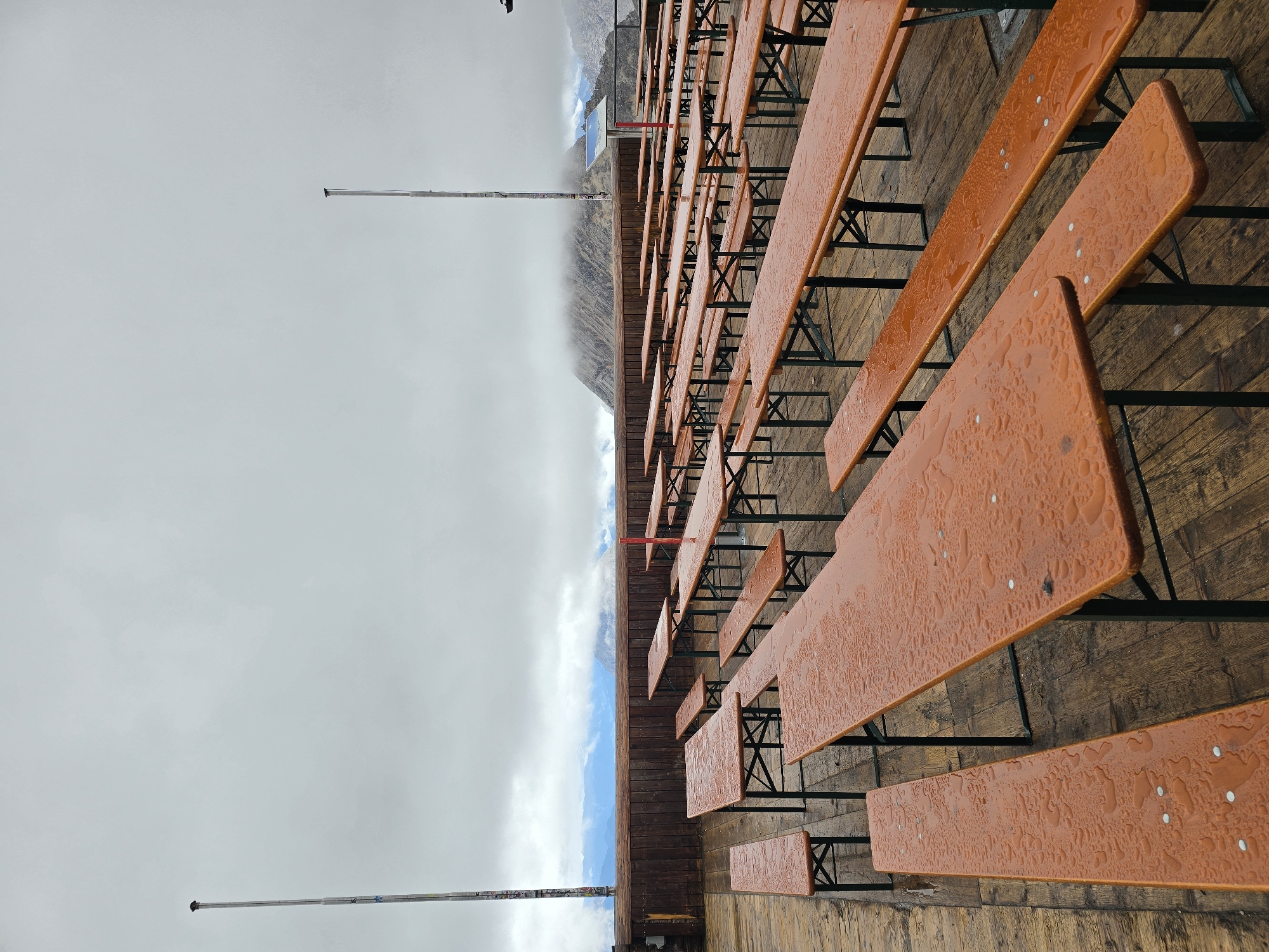 The image depicts an outdoor seating area, likely a beer garden, at a high-altitude location. The scene is dominated by rows of empty, orange-topped picnic tables and benches, many of which are wet with raindrops. The tables are arranged on a wooden deck, which shows signs of wear and tear. In the background, a wooden railing separates the seating area from a view of misty mountains under a cloudy, overcast sky. Two tall flagpoles are visible, one to the left and one to the right of the frame. The overall impression is one of a quiet, possibly abandoned, outdoor venue experiencing inclement weather. While the specific location in Garmisch-Partenkirchen, Germany, is not explicitly identifiable from the visual elements alone, the style of picnic tables and the mountainous landscape are consistent with alpine regions often found near such Bavarian towns.