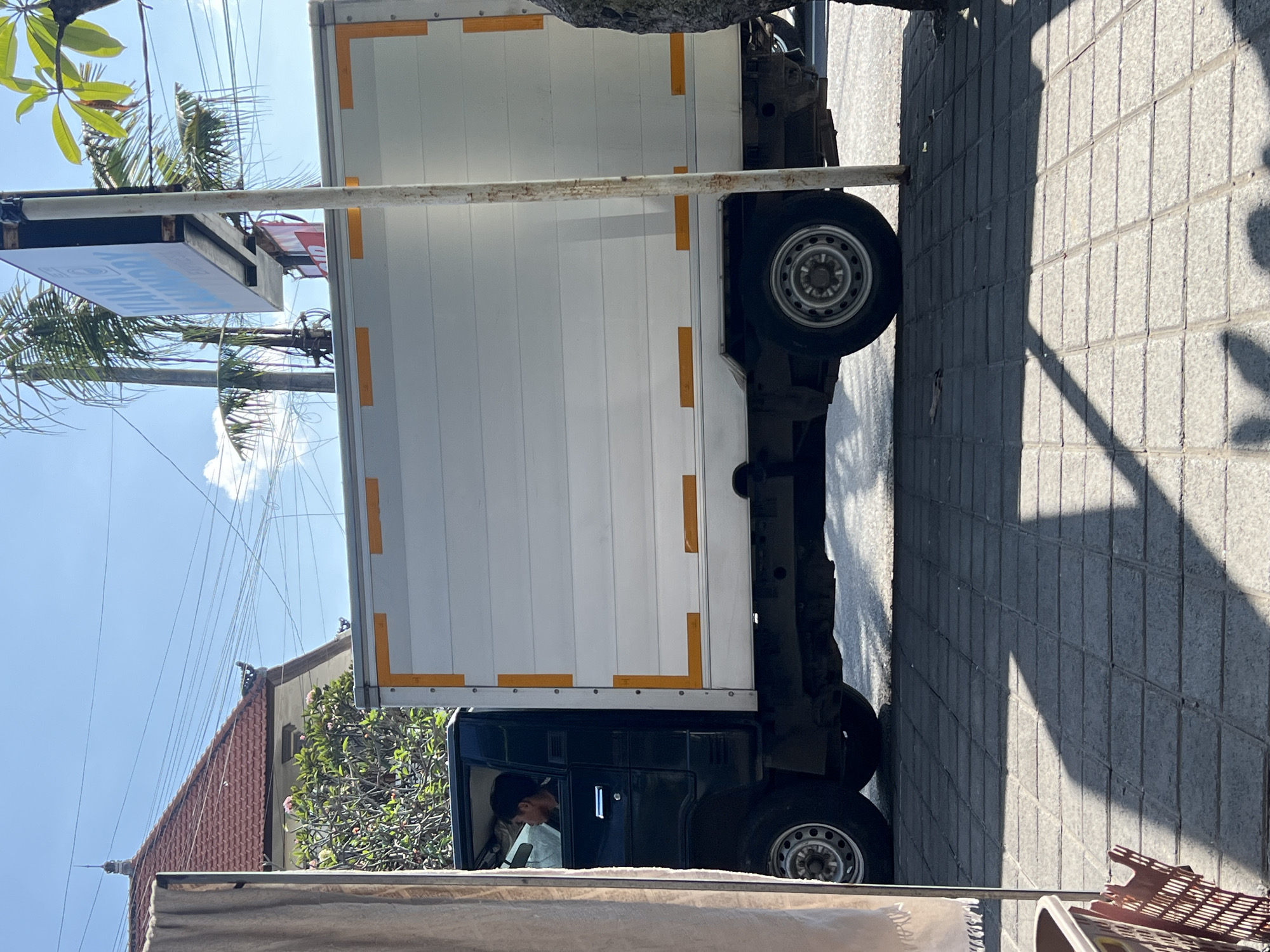 The image captures a side view of a white box truck with yellow reflective stripes on its side, parked on a tiled sidewalk in Denpasar, Indonesia. The truck is dark green on the front cab, and a man is visible inside, looking towards the right. To the left of the truck, a beige curtain hangs, and a brown plastic basket is partially visible in the lower left corner. In the background, a red-tiled roof house with a traditional Balinese architectural style and lush green foliage can be seen. A pole with a "VIANA LAUNDRY" sign is situated to the right of the truck. The scene is bathed in bright sunlight, casting shadows on the sidewalk.