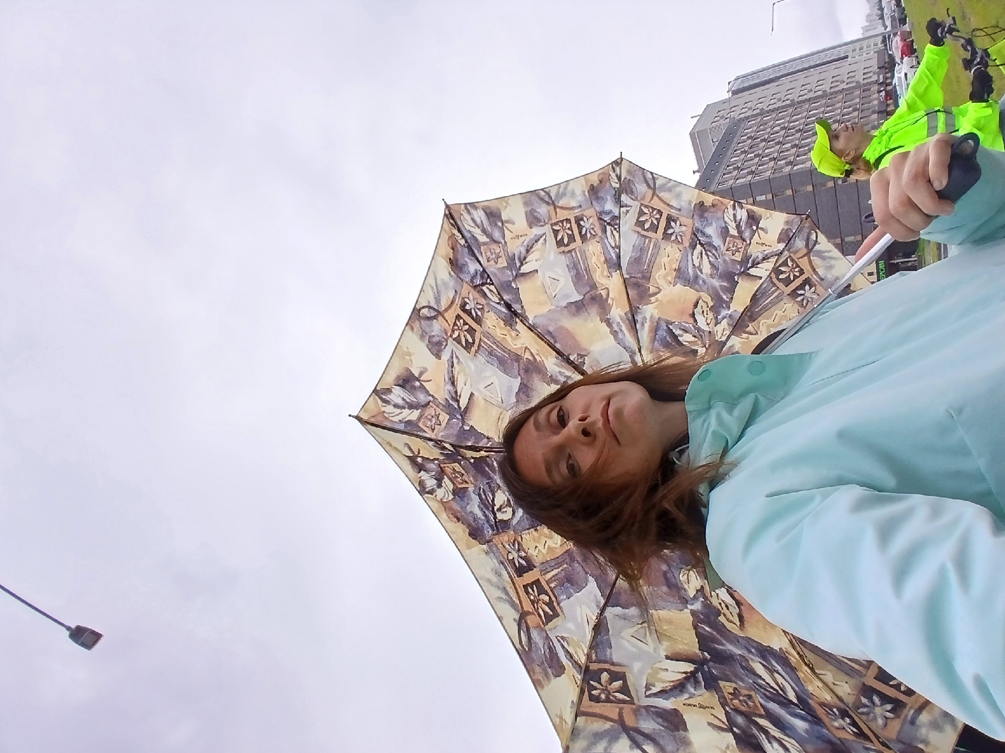 A low-angle, wide-shot captures a woman holding a patterned umbrella outdoors, with a cyclist in a neon-green jacket and cap visible in the background. The woman, positioned in the foreground, is looking slightly upwards and to her left, her brown hair windswept. She wears a light blue jacket. The umbrella, held by its handle in her right hand, is open and displays a repeating pattern of floral motifs within square frames, interspersed with abstract designs. Behind her, the cyclist, also facing left, is on a bicycle. The environment is urban, with a tall, modern building with many windows dominating the background. Cars are visible on a road to the right of the cyclist and building. The sky is overcast, suggesting a cloudy or possibly rainy day. A street lamp is visible in the upper left portion of the frame. The scene appears to be Yekaterinburg, Russia, given the architectural style of the building and the general urban setting.