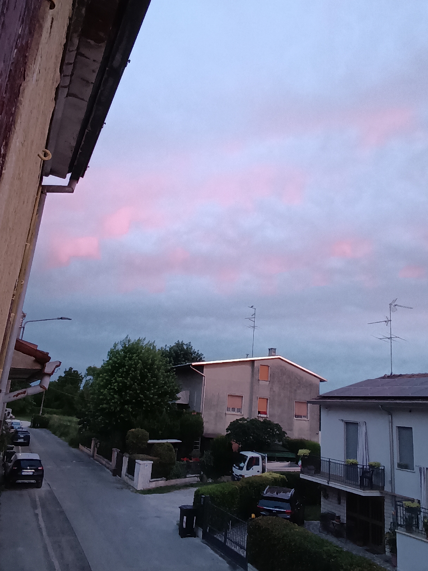 The image captures a street scene in Viadana, Italy, during twilight. The sky is a dusky blue with streaks of pink clouds. On the left side of the frame, a building's textured, beige exterior wall and a portion of its gutter are visible. A street light extends over the road.
The street itself is paved and curves slightly to the right. Several cars are parked along the road: a black car, a dark grey SUV, and another dark vehicle further up the street. A white van is parked in a driveway to the right.
Several houses are present. One house in the mid-ground has a light-colored facade, with several windows and what appears to be an attic window. A large, dense green tree is located in front of this house. To the right, a white house with a balcony is visible, with potted plants on the balcony and solar panels on the roof. Another building, potentially a garage or an extension of the white house, is partially visible on the right edge of the frame.
The foreground and the right side of the image are bordered by hedges and fencing, suggesting residential properties. Two television antennas are prominent on the roofs of some buildings. The overall impression is a quiet suburban or residential street as daylight fades.