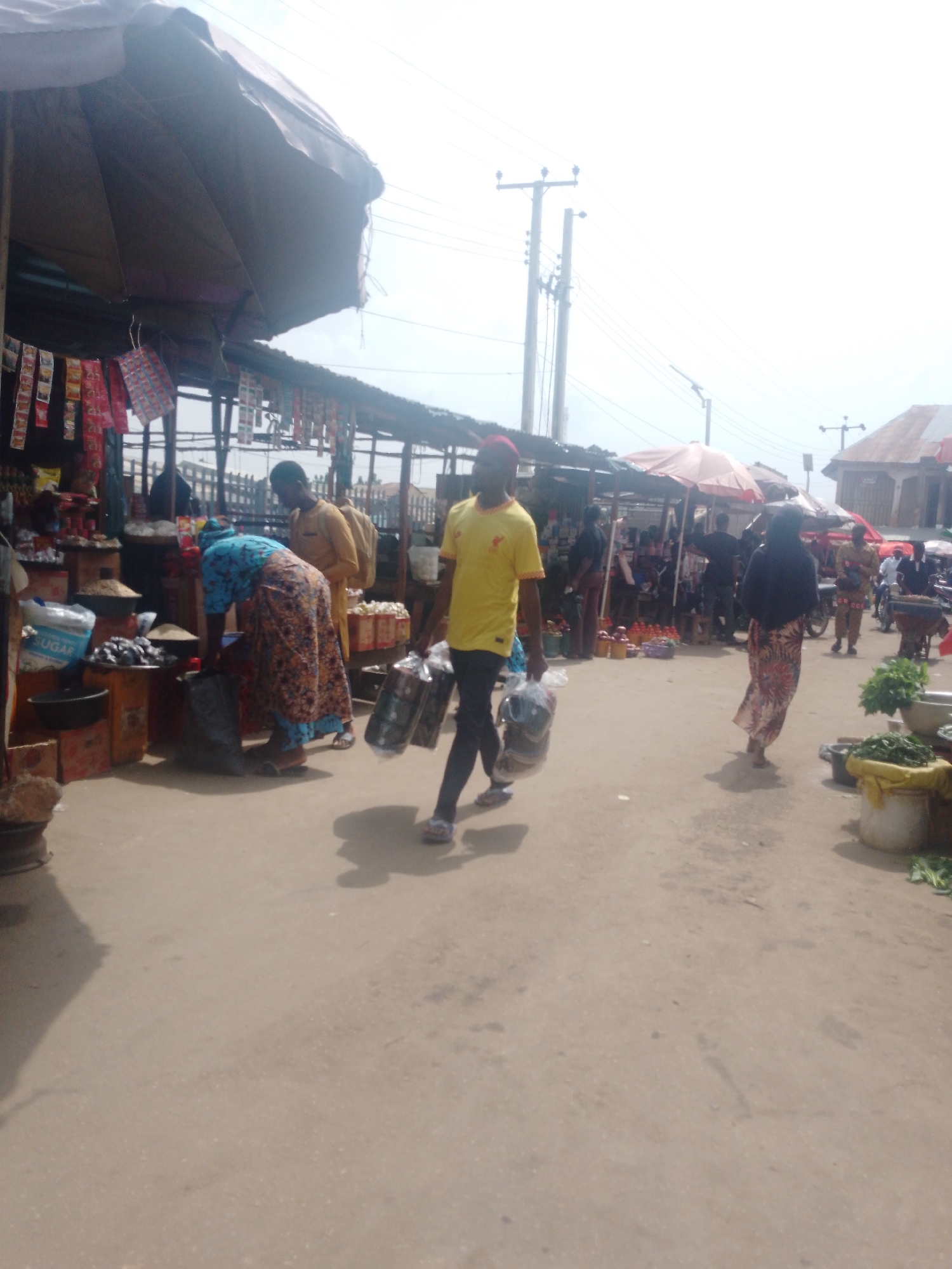 The image depicts a bustling marketplace in an unidentified city. The foreground is dominated by a sandy ground with visible shadows. In the middle ground, a man wearing a yellow shirt and dark pants carries two bags of groceries. To his left, a woman in a blue and floral patterned dress shops at a stall. Several other individuals are present in the background. Stalls lined with various goods and covered by awnings are also visible. Power lines can be seen overhead, adding to the urban context. The sky appears overcast.