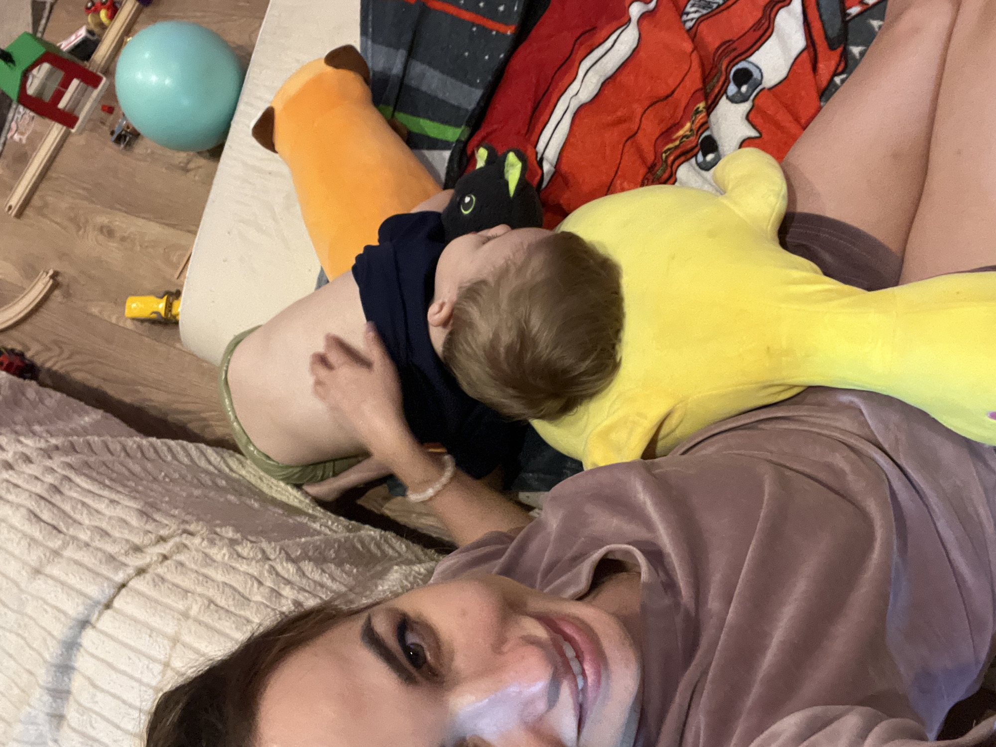 An overhead medium shot shows a woman lying on a bed next to a child. The woman is on the left and takes up the left half of the frame. She is smiling and looking at the camera. She has long, dark hair pulled back and is wearing a mauve, v-neck, short-sleeved shirt. Her right arm is bent and her hand is resting on her leg. The child is on the right, asleep, with their head on a large yellow stuffed toy. The child is wearing a dark blue shirt and light green shorts. They are lying on an orange stuffed toy that resembles a dog. Behind the child and to the right is a red blanket with a cartoon character design. The background shows a wooden floor with toys scattered on it, including a wooden train track, a green toy building, and a blue ball. The overall impression is that of a casual, indoor scene.