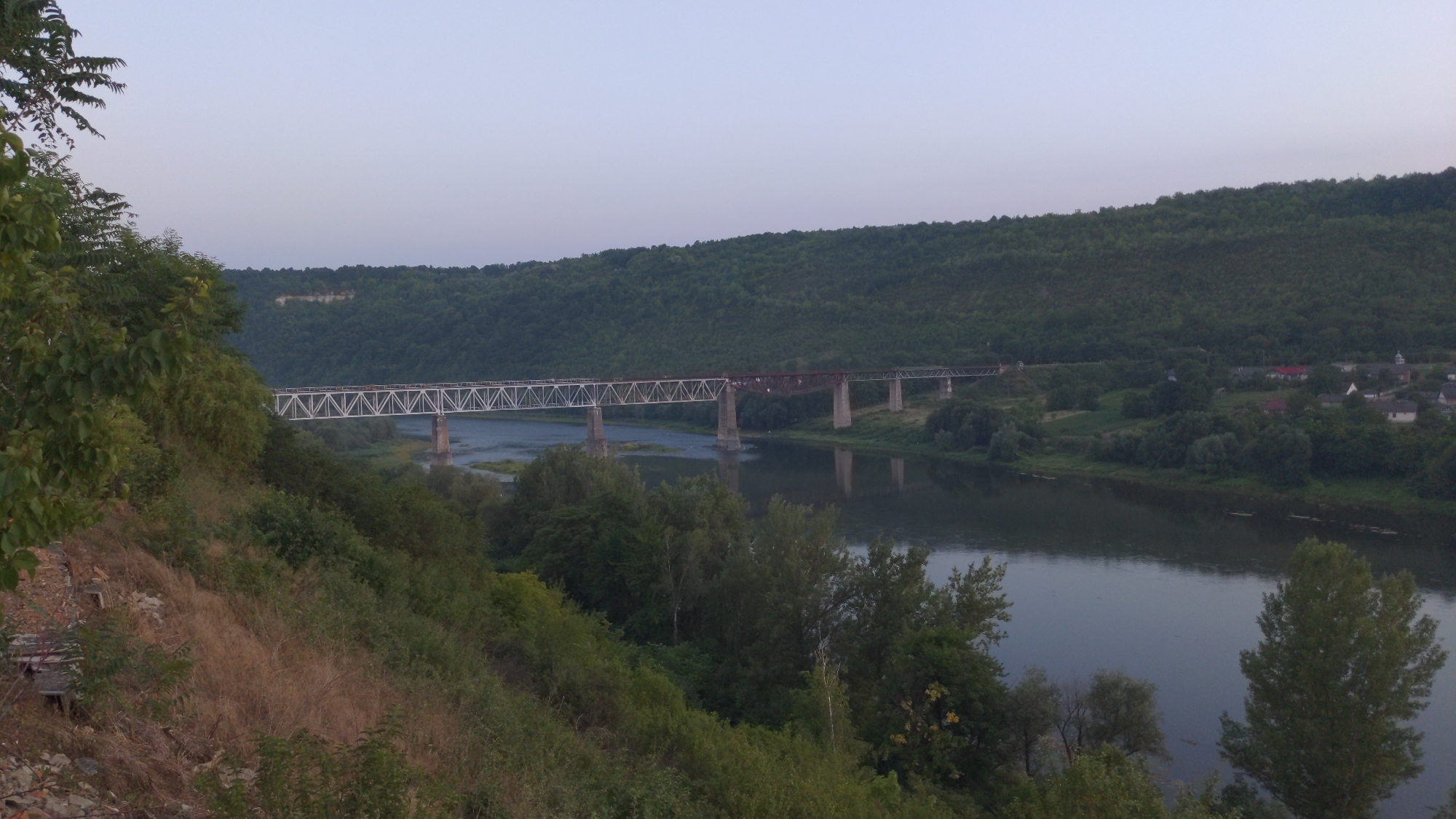 The image depicts a panoramic view of a river valley with a railway bridge spanning across it. The bridge is a prominent structure, characterized by its metal truss design. On the far side of the river, a lush green forested hill rises, with a small village nestled at its base. The village features several houses with red and white roofs, and a church with a steeple is visible. The river itself is calm and reflects the surrounding landscape. The foreground of the image shows a steep, overgrown slope covered with vegetation and dry grass. The sky is a pale, hazy blue, suggesting either early morning or late evening. The scene appears to be a rural landscape, likely in Zalishchyky, Ukraine, given the distinctive S-shaped bend of the Dniester River that characterizes the area. There are no people visible in the image, and the focus is on the natural scenery and infrastructure.