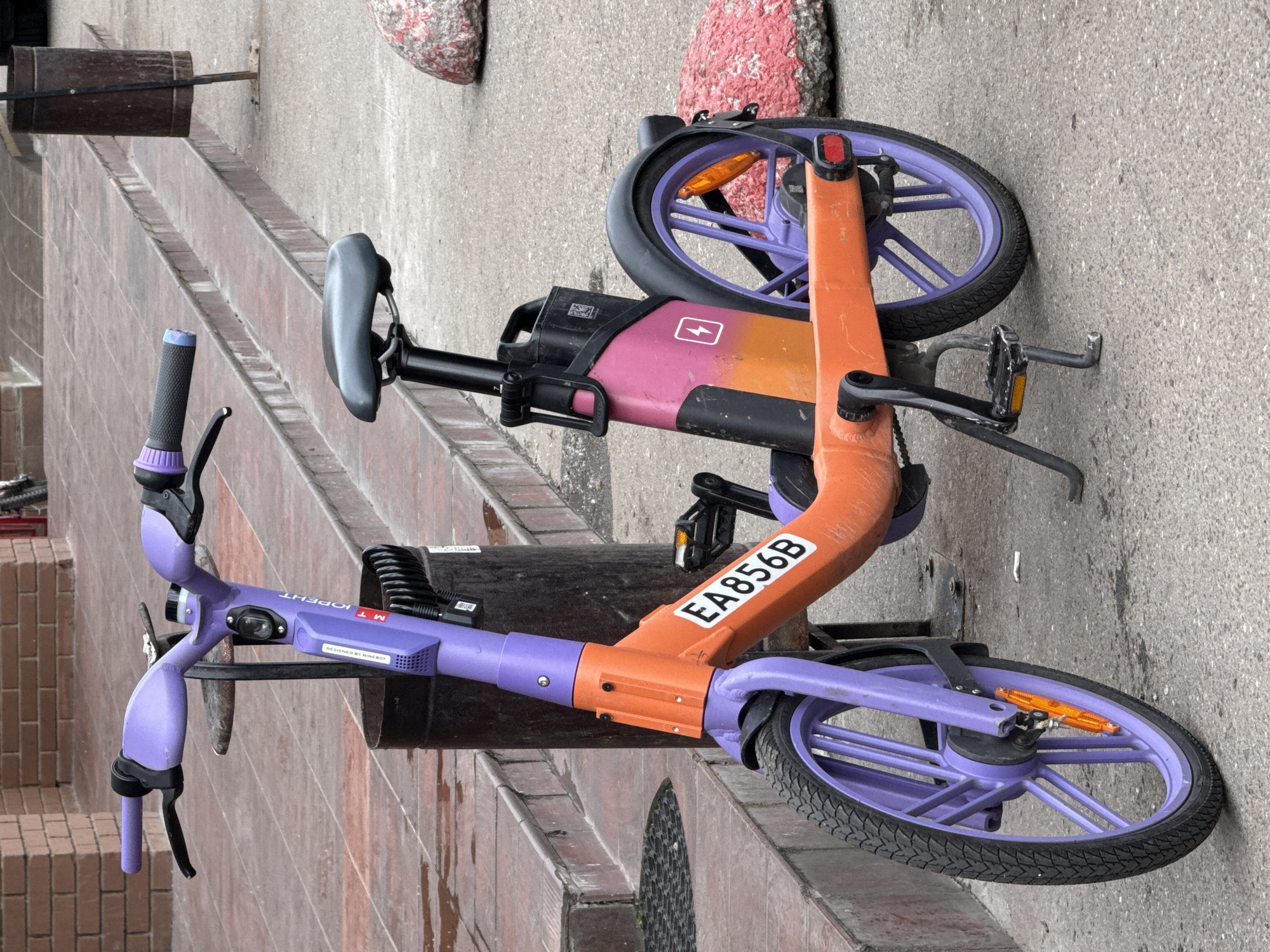 A purple and orange electric scooter is parked on a paved sidewalk next to a set of brick stairs and a trash can. The scooter has a black seat, purple handlebars, and a license plate with the characters EA856B. The scooter is upright, supported by its kickstand. There are two large pink and red rocks on the sidewalk to the right of the scooter. The background shows a brick building and more stairs. The lighting suggests it is daytime.