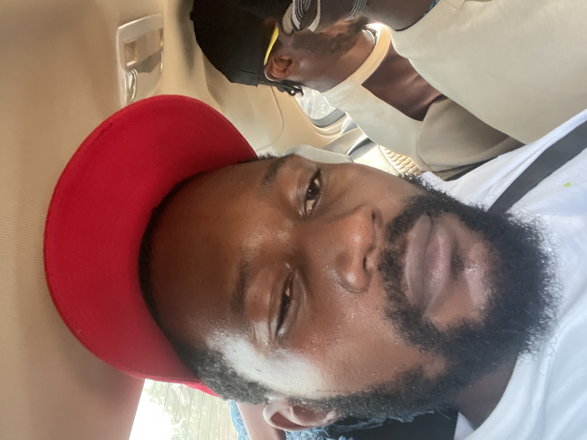 The image is a close-up, rotated view, featuring a man inside what appears to be a vehicle. The main subject, prominent in the foreground, is a man with dark skin, a full black beard, and short dark hair. He is wearing a white shirt and a red baseball cap, worn backward, and looks directly towards the camera with a neutral expression. His skin has a visible sheen, suggesting warmth or humidity.
In the background, another person with dark skin is partially visible. This individual is wearing a light-colored top, possibly cream or light beige, and a dark cap or head covering. Yellow sunglasses or a yellow strap are visible near their ear.
The setting is the interior of a vehicle, indicated by the light-colored (beige/cream) headliner and trim, along with a silver handle or hook seen in the upper left. Through a window, glimpses of green foliage are visible, suggesting it is daytime. The casual attire and vehicle interior point to a moment during travel or a commute. The location context indicates this scene takes place in Abuja, Nigeria. There is no visible text in the image.