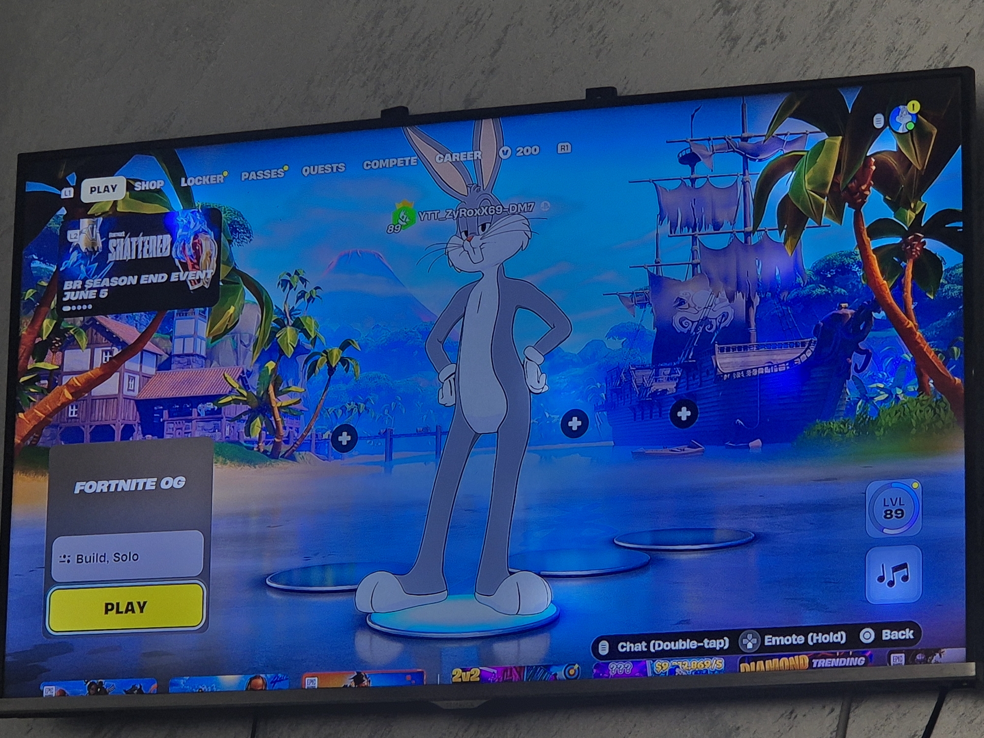 The image shows a television screen displaying the game "Fortnite." The main screen features Bugs Bunny as a character, standing on a platform in front of a beach scene with a pirate ship. The player's username, "YTT_ZyROxX69-DM7," is visible above the character. On the left side of the screen, there's a "Fortnite OG" game mode selection highlighted, with options to "Build, Solo" and a yellow "Play" button. The player's level, "LVL 89," is displayed on the right side of the screen. Various menu options are visible at the top and bottom of the screen, including "Play," "Shop," "Locker," "Passes," "Quests," "Compete," "Career," "Chat," "Emote," and "Back." An event banner for "Shattered BR Season End Event" is shown in the top left corner.
