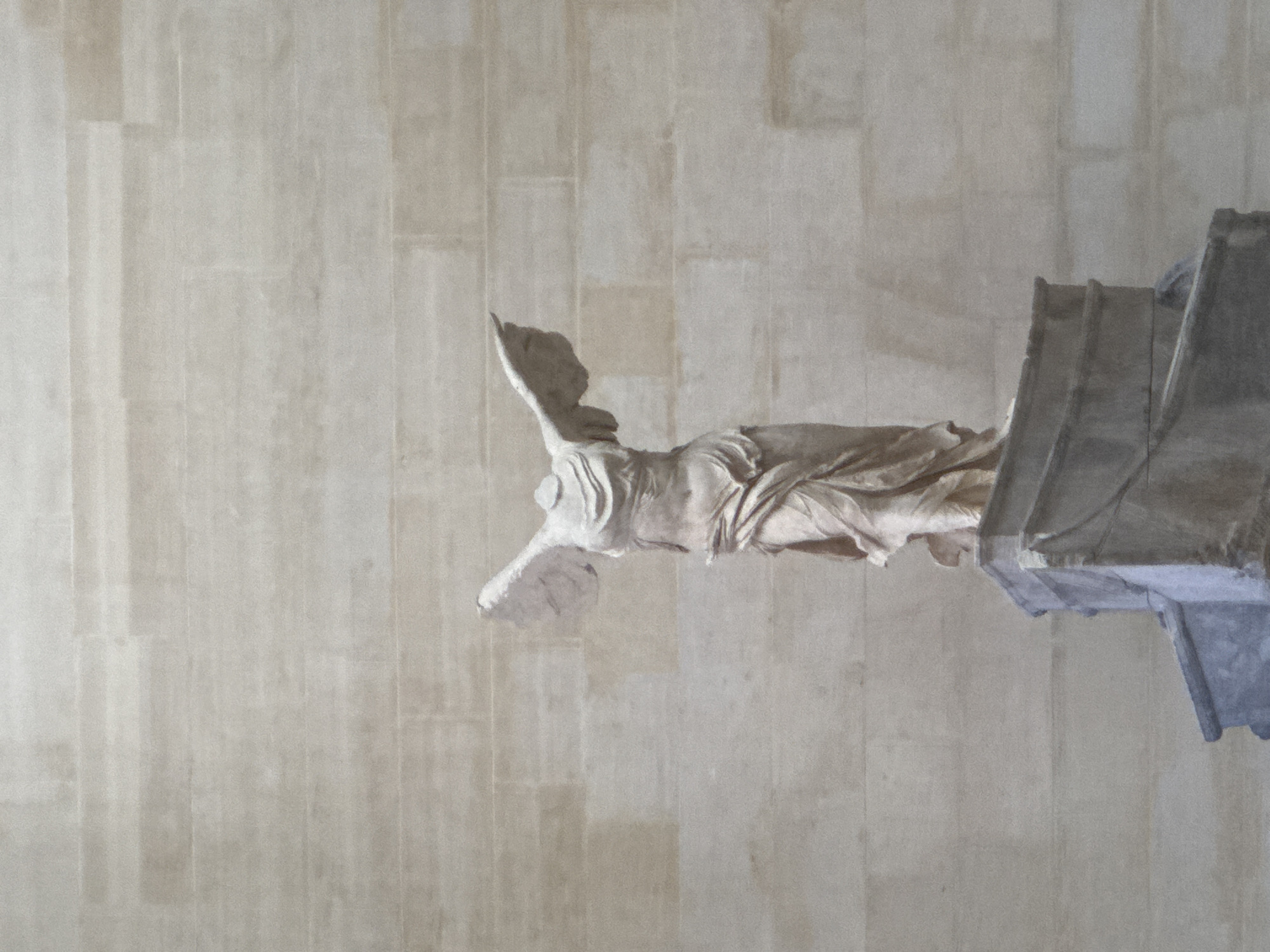 The image depicts the Winged Victory of Samothrace, a marble sculpture located in the museum of the Louvre in Paris, France. The headless statue, dating back to the Hellenistic period, stands proudly on a pedestal against a textured, stone-like wall. The figure is depicted with its wings outstretched and a flowing garment draped around its body, suggesting motion and triumph. The lighting is even, highlighting the detail and form of the sculpture. The overall scene is one of historical and artistic significance, captured in a static, observational manner.