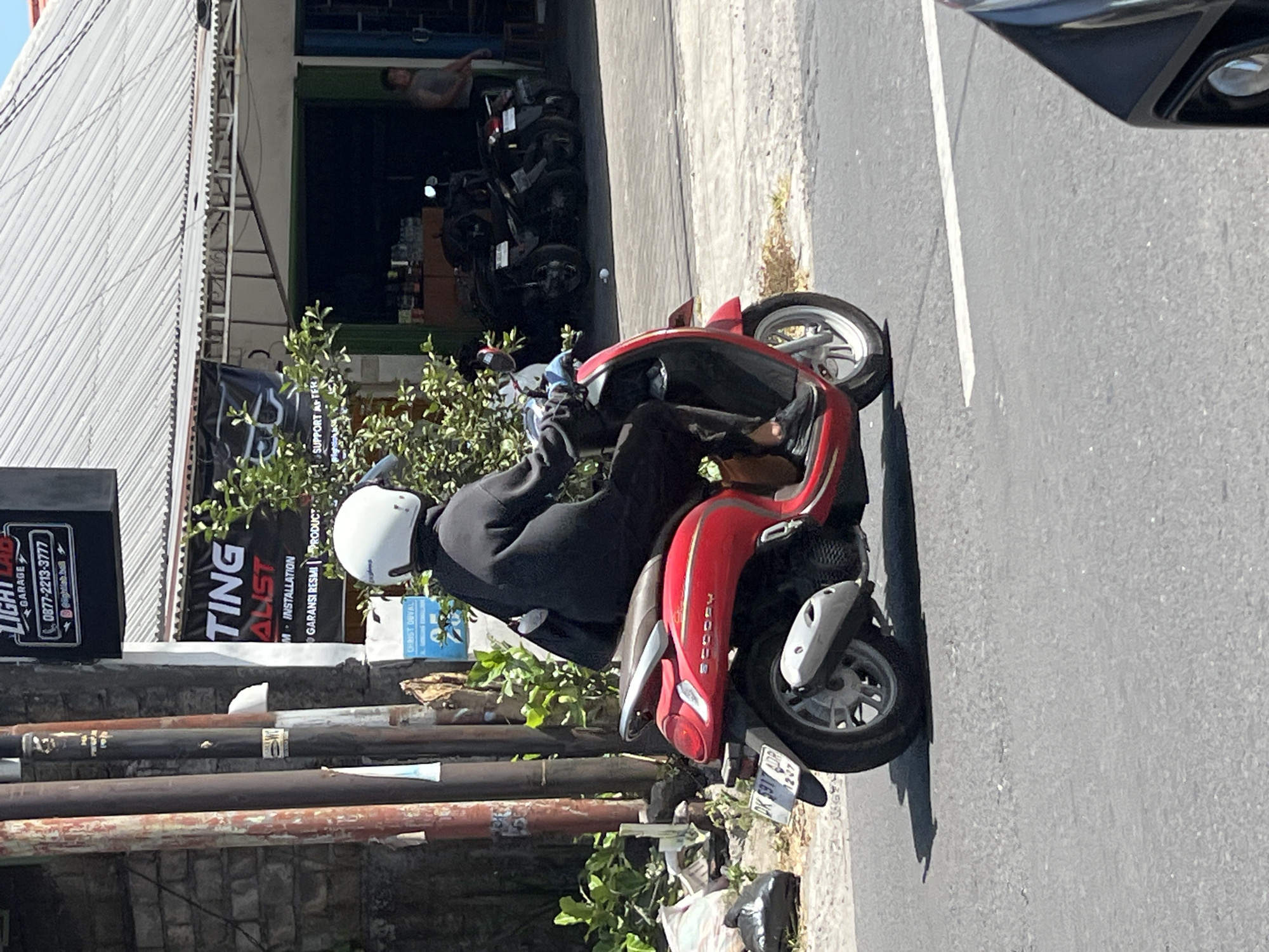 A person wearing a black outfit and a white helmet is riding a red scooter on a street in Denpasar, Indonesia. The scooter has a license plate that reads "DK 5917 ADR". In the background, there are buildings with corrugated metal roofs, a store with motorcycles parked outside, and a person standing in the doorway. The street is paved with asphalt and has a white line marking a lane. There are some plants and trash bags on the side of the road. The image appears to be taken during the daytime, with bright sunlight casting shadows.