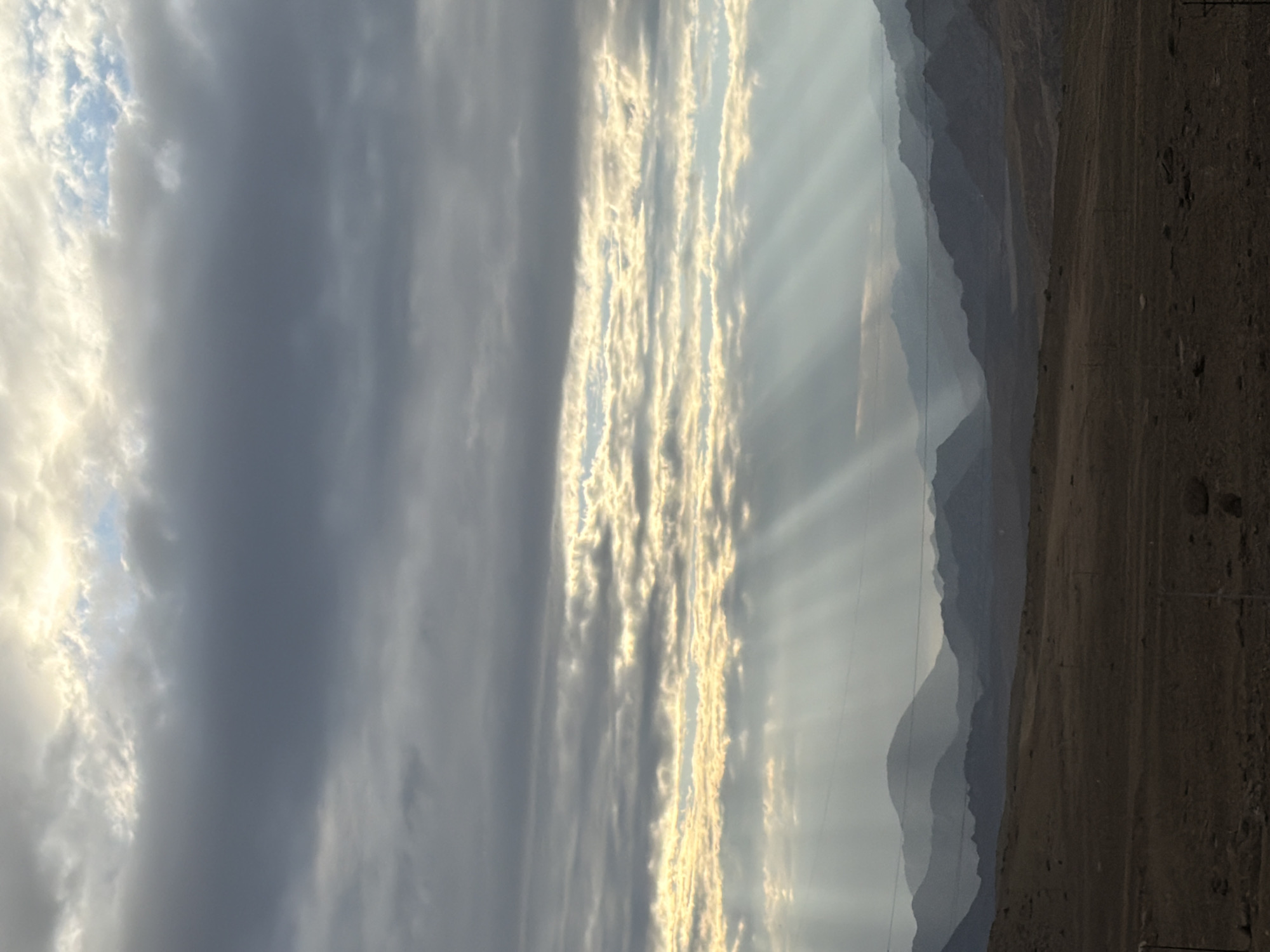 The image shows a panoramic view of a mountain range under a dramatic sky filled with clouds and sunbeams. The foreground is a desolate, dry landscape of brown earth with scattered rocks. In the middle ground, rolling hills and more distant mountains are silhouetted against the hazy sky. The sky is characterized by layers of dark and light clouds, with bright rays of sunlight piercing through them, creating a dramatic effect. There are no people or specific structures visible in the image, suggesting a remote, natural environment. The scene evokes a sense of vastness and natural beauty.