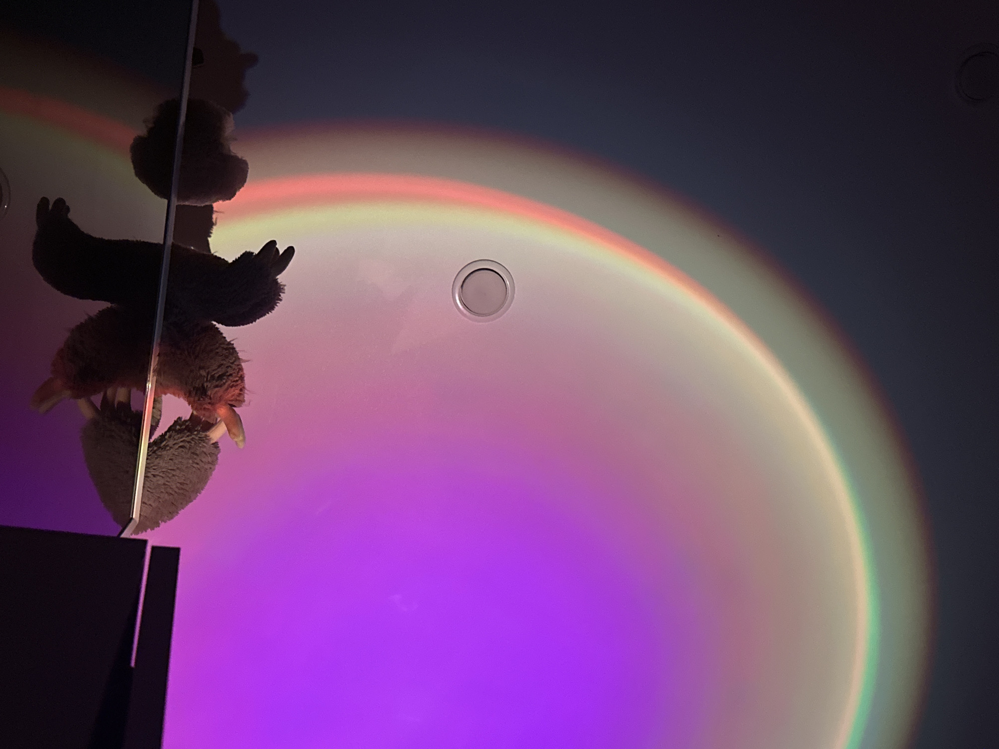 This close-up image presents an indoor scene, likely a room in Chaikovsky, Russia, characterized by atmospheric lighting. The dominant feature is a large, semi-circular light projection on a wall or ceiling, creating a radiant gradient of colors. The projection transitions from a vivid purple/magenta at the bottom, through soft pinks, yellows, and greens, to a muted greyish-blue towards the top, resembling a celestial or sunset effect. The overall dimness suggests it is evening or night.
In the foreground, on the left side, a stuffed animal, appearing to be a sloth or similar furry creature, hangs upside down. Its body is largely silhouetted against the bright projection, but details of its textured fur and distinct claws are visible. It appears to be suspended from a thin, vertical support, possibly a pole or part of an unseen fixture. Below the stuffed animal, in the bottom-left corner, a dark, rectangular object, possibly a monitor or speaker, is partially visible.
Embedded in the surface where the light is projected, a small, round, silver fixture, likely a recessed light or sensor, is visible near the center of the color arc. A faint, circular mark is also discernible in the darker upper-right portion of the surface. The scene conveys a calm and artistic ambiance. There is no visible text in the image.