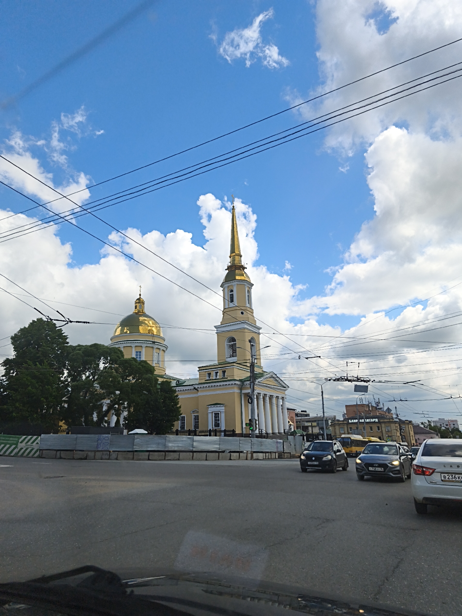 The image presents a daytime street scene in Izhevsk, Russia, dominated by the majestic St. Michael's Cathedral. The cathedral is a large, yellow-painted building with prominent white neoclassical columns at its base, leading up to a golden dome and a tall, elegant golden-spired bell tower, each topped with a cross. Lush green trees partially obscure the cathedral's left side.
The view is from inside a vehicle, with reflections visible on the windshield. On the road, several cars are present: a black Suzuki SX4, a gray Hyundai Solaris, and a white Lada Granta in the foreground. A yellow bus is visible further down the street. Russian license plates with the region code "18" (Udmurt Republic), such as "H692XT 18", "C748YY 18", and "B236KO 18", are clearly seen on the vehicles, confirming the location.
The sky is a vivid blue with numerous large, white cumulus clouds. A complex network of black power lines and trolleybus wires crisscrosses the upper part of the image. To the left of the road, a construction barrier consisting of metal fencing and concrete blocks is in place. Some indistinct, graffiti-like text is visible on the metal fence. In the background, beyond the road and partially obscured by the wires, another building features a sign that reads "БАНК АВАНГАРД" (Bank Avangard). The scene portrays a typical urban environment under clear, bright weather conditions.