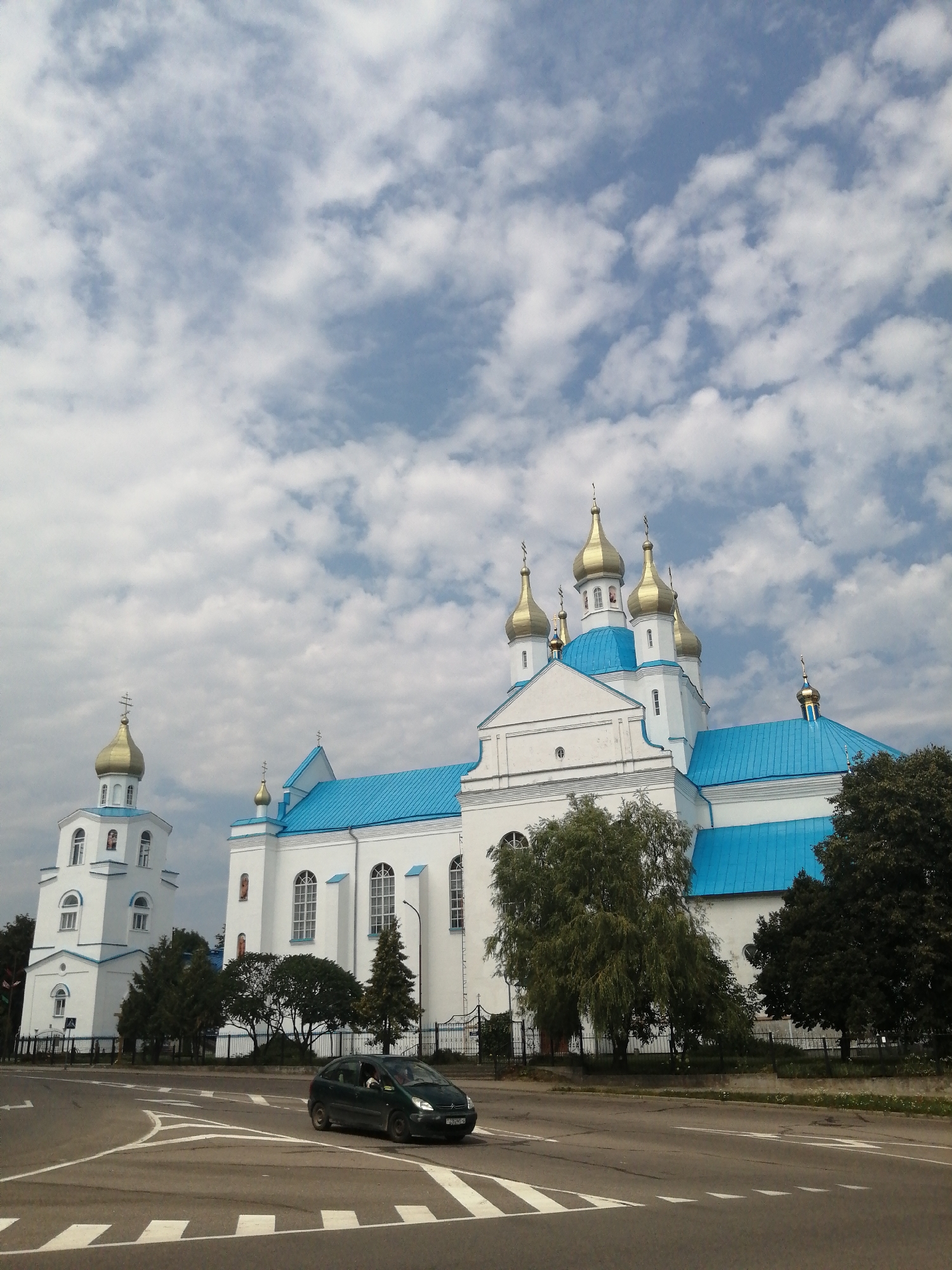 In Slonim, Belarus, a wide, paved road with white lane markings is visible in the foreground, with a dark-colored car turning right. Behind the road, a large, white building with a bright blue roof and golden domes stands prominently. To the left of this building is a smaller, white bell tower, also with a golden dome. Trees and a dark metal fence are situated between the building and the road. The sky is partially cloudy with patches of blue. The architectural style suggests a religious structure, likely a church or cathedral.