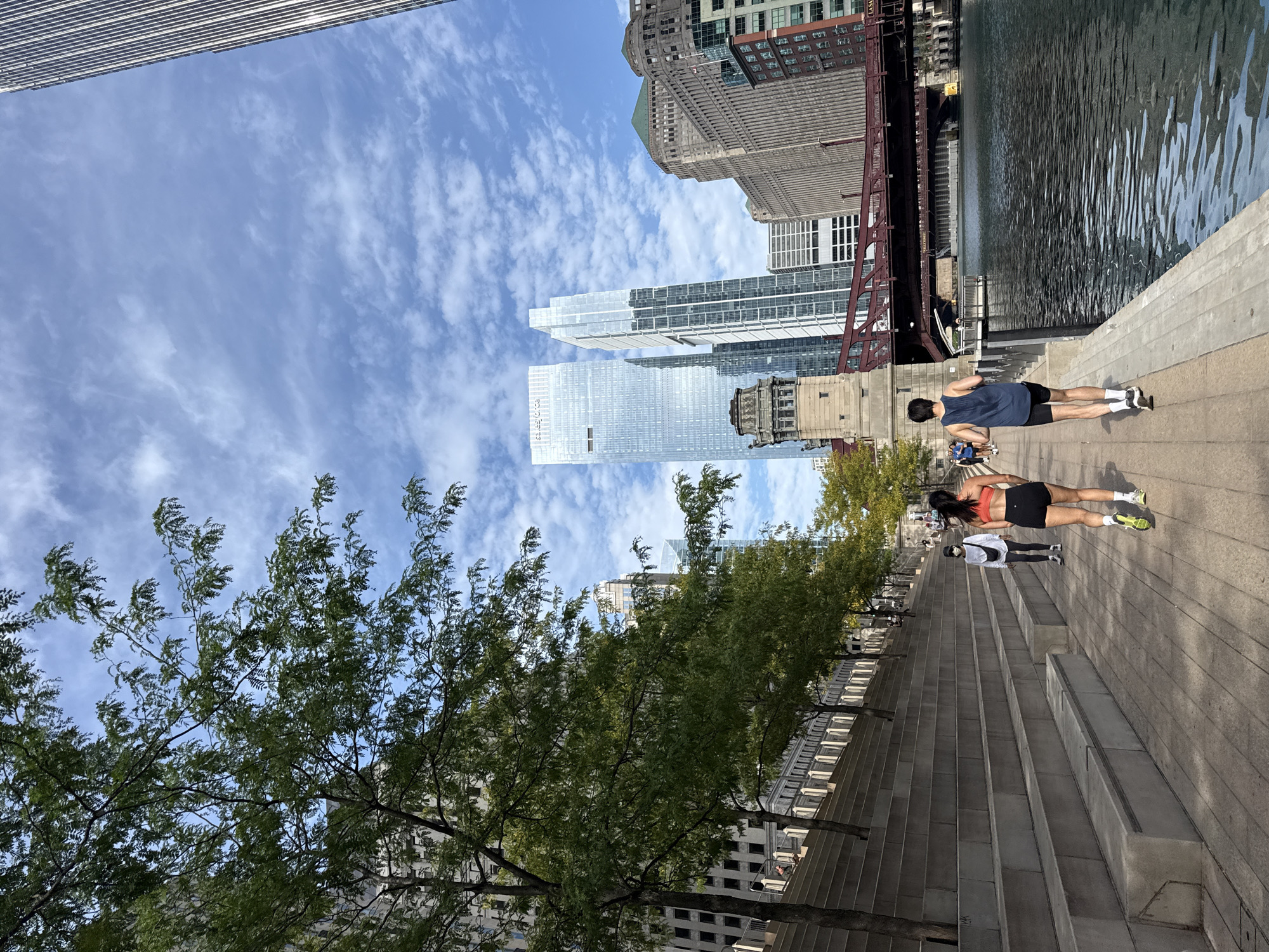 An eye-level, full shot captures a man and a woman jogging on a paved riverwalk in Chicago, Illinois, USA, on a cloudy day. The man, wearing a dark blue tank top and black shorts, runs ahead of the woman, who wears a red sports bra and black shorts. The riverwalk is lined with tiered concrete seating on the left and the Chicago River on the right. In the background, skyscrapers tower over a red bridge, and trees with green leaves are visible. Several other people are walking and jogging in the distance on the riverwalk and on the bridge. The sky is blue with scattered white clouds.
