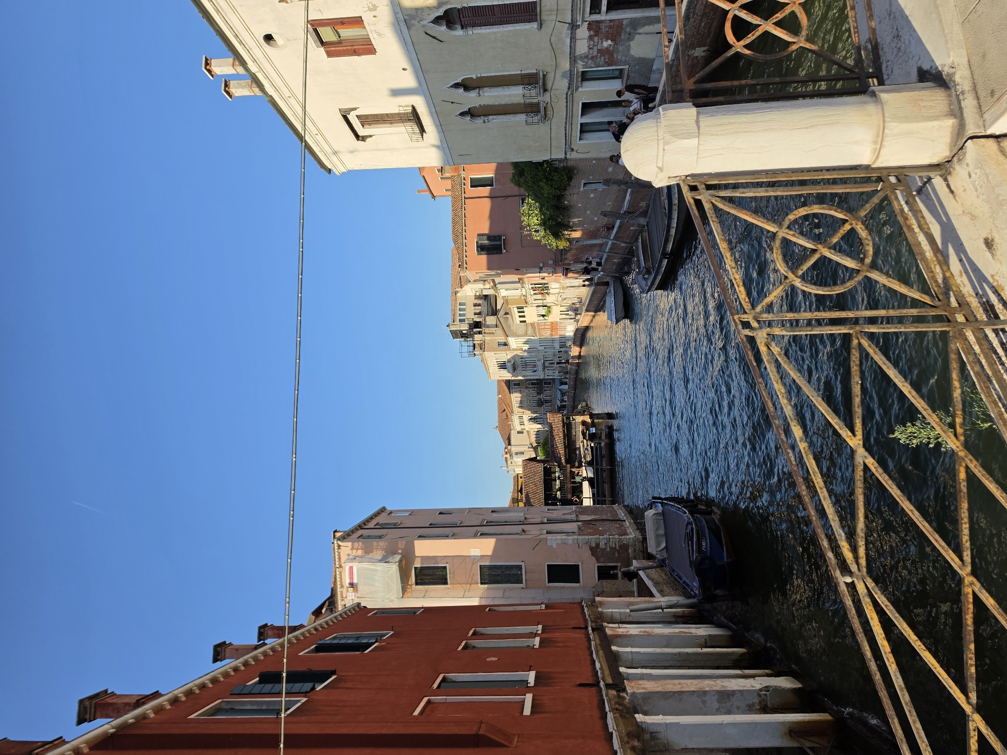 This is a photograph taken from a bridge overlooking a narrow canal in Venice, Italy. The canal is lined with colorful, multi-story buildings with traditional Venetian architecture. On the left, a vibrant red building features arched openings at its base and shuttered windows. To the right of the canal, buildings of lighter hues, including pale yellow and pink, display arched windows and balconies. Several boats are moored along the canal banks, including a dark-hulled boat with a grey cover in the foreground. In the distance, more buildings create a picturesque urban landscape, with a clear blue sky overhead. A rusty metal railing with circular decorative elements runs along the bridge in the foreground. A white stone pillar stands prominently on the bridge. Several people are visible on the walkways beside the canal.