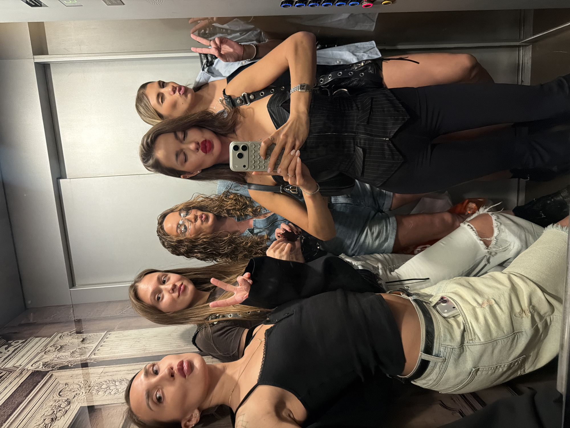 A group of five young adult women are observed posing for a mirror selfie inside an elevator located in Khimki, Russia. The elevator interior features light-colored walls; the left wall displays a sepia-toned mural depicting classical architectural elements, including fluted columns with Corinthian capitals. The right wall contains a vertical control panel with illuminated buttons, showing blue, red, and green lights.
From left to right, the women are positioned as follows:
1. A woman with long, dark brown hair, wearing a black spaghetti strap top with lace trim, light-colored distressed jeans with a dark belt, and a gold necklace. She is leaning against the wall, striking a pose with pouted lips and eyes slightly open, looking towards the right. A silver smartphone is partially visible in her right rear pocket.
2. A woman with long, straight light brown hair, wearing a dark t-shirt and light-colored ripped jeans. She holds her right hand up, displaying a peace sign. Her face shows a slight smirk.
3. Partially obscured, a woman with curly brown hair, wearing clear-framed glasses and a light-blue denim jumpsuit. Her lips are pouted, and she holds dark sunglasses in her left hand.
4. At the center, a woman with long, dark brown hair, wearing a black pinstriped corset-style top with a front zipper and black pants. She holds a silver smartphone with a clear, ridged case in both hands, positioned to capture the reflection. Her eyes are closed, and her red-lipsticked lips are pouted. She wears a watch on her left wrist and a black studded shoulder bag.
5. On the far right, partially reflected, a woman with blonde hair, wearing a light blue collared shirt. She forms a peace sign with her raised left hand, winks, and pouts.
The women are engaged in taking a group photograph, with multiple individuals making peace signs and pouting expressions.