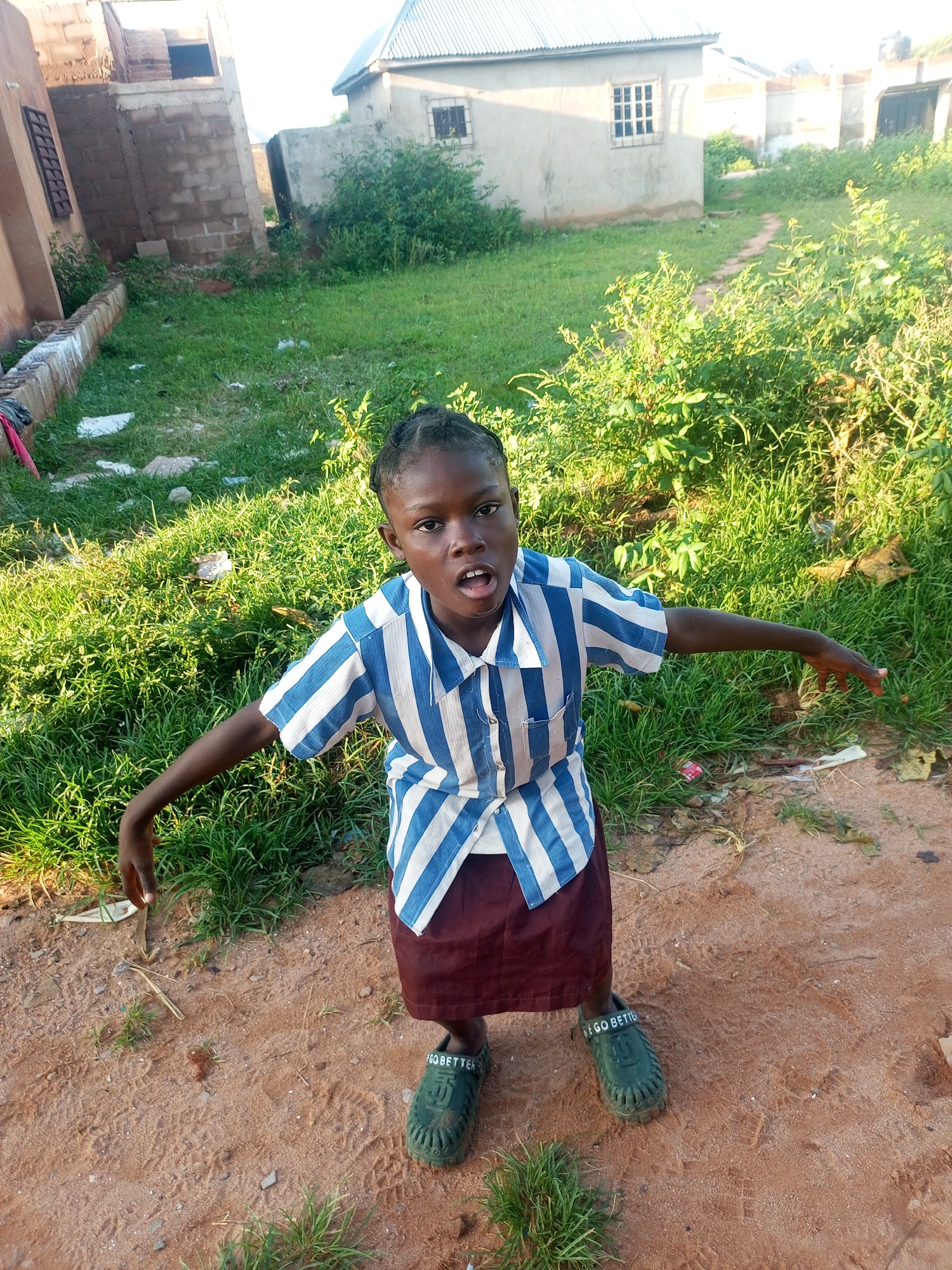A young Black girl stands on sandy ground in an outdoor setting, possibly Tempure, Nigeria. She is wearing a blue and white striped shirt and a maroon skirt. Her arms are outstretched, and her mouth is open, as if she is speaking or shouting. Green clogs are on her feet. The background shows a grassy area with bushes and a building under construction. The lighting suggests it is daytime, with sunlight illuminating the scene.
