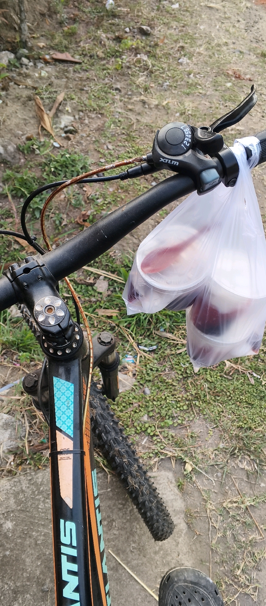 A close-up, vertical shot captures the handlebars and front portion of a bicycle, with a plastic bag containing two take-out containers hanging from the left handlebar. The containers appear to hold a reddish-colored food or drink, and one has a spoon visible. The bicycle's handlebars are black, and the gear shifter is visible on the right side, labeled "XTLM" and with numbers indicating gears. The bicycle frame is black and orange, with some text and a blue patterned sticker visible on the downtube. The foreground shows the front tire of the bicycle, and a black shoe is visible at the bottom right. The background consists of dry, patchy grass and dirt, with some scattered debris, suggesting an outdoor, possibly rural or undeveloped area. There are no people visible in the frame. The scene is forensic in its detail and matter-of-fact presentation.